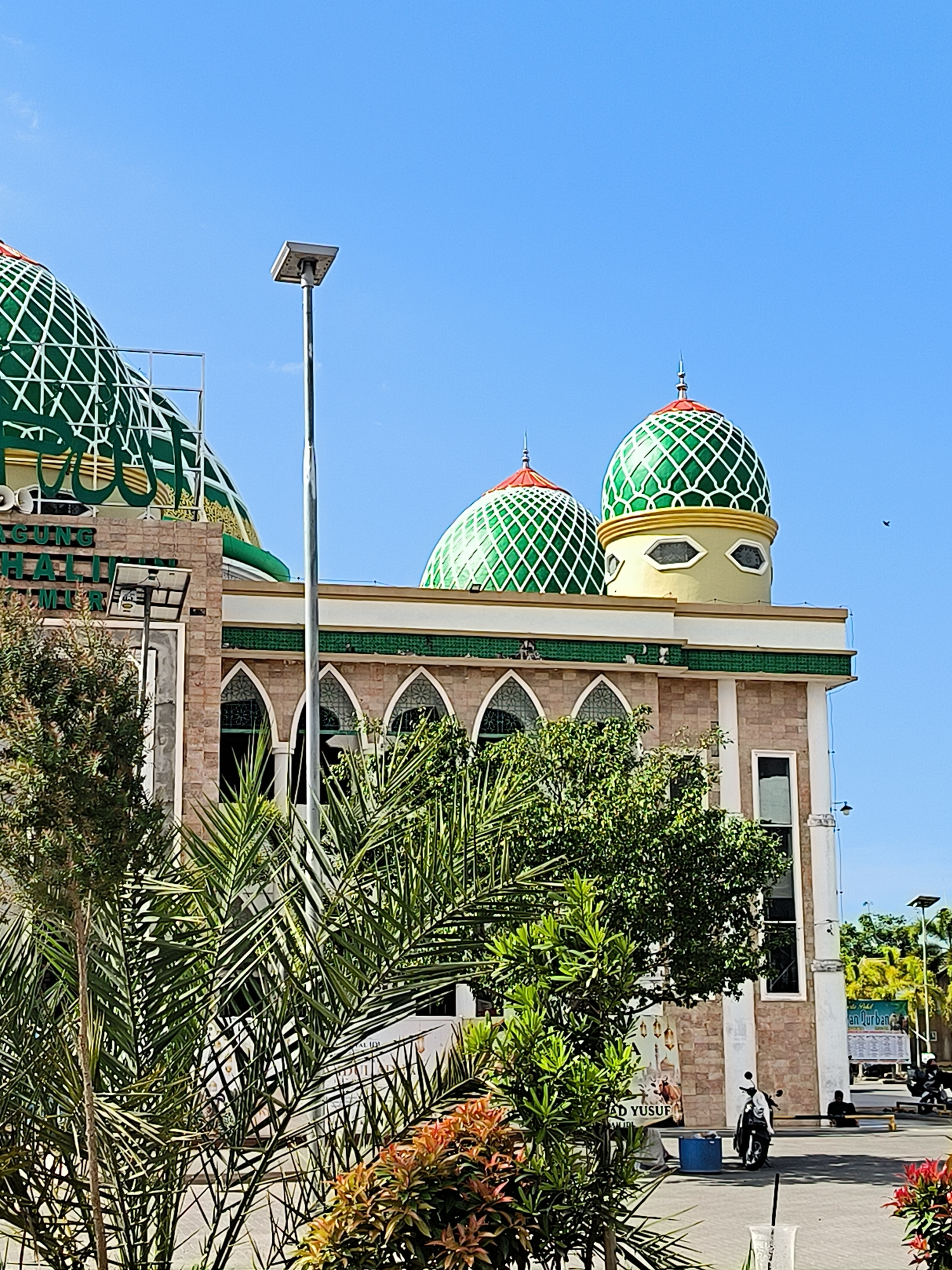 The image captures a section of a mosque in Titibaro, Indonesia, on a bright day with a clear blue sky. The mosque features distinct architecture, including multiple domes with green and white geometric patterns, topped with red accents. The building is constructed from light brown brick with green and white trim. Arched windows are visible on the side.
In the foreground, a variety of greenery including shrubs and palm trees partially obscure the view of the mosque. A tall metal pole stands near the building.
In the background, further details of the city are visible, including buildings, trees, and vehicles. Two people are visible to the right side, sitting on the ground. A billboard is visible in the distance, as well as a motorcycle.