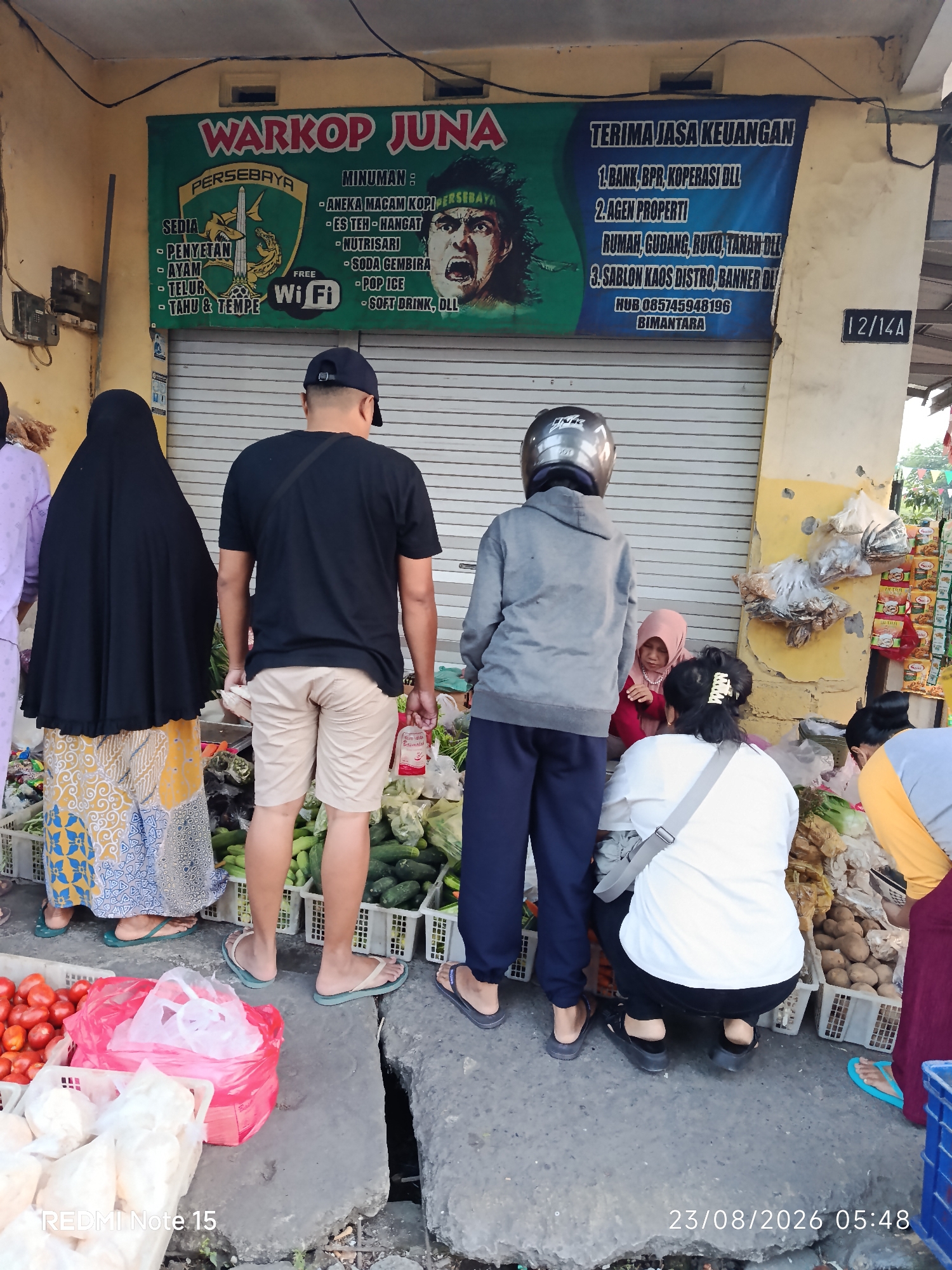 The image depicts a street scene in Keboansikep, Indonesia, likely in front of a small shop or warung. A green banner, "Warkop Juna," is prominently displayed, advertising food and drinks, and also offering financial services such as loans, property services, and custom printing. Several individuals are present. A woman in a black hijab and patterned skirt stands by a stall filled with vegetables. Beside her, a man wearing a black t-shirt and light-colored shorts is seen from the back. Further to the right, two people wearing helmets are interacting with a child in a pink hijab. The foreground features stalls displaying a variety of produce, including tomatoes, cucumbers, and potatoes, along with plastic bags of goods. The setting appears to be an outdoor market or a roadside vendor area. The timestamp "23/08/2026 05:48" is visible in the lower right corner.