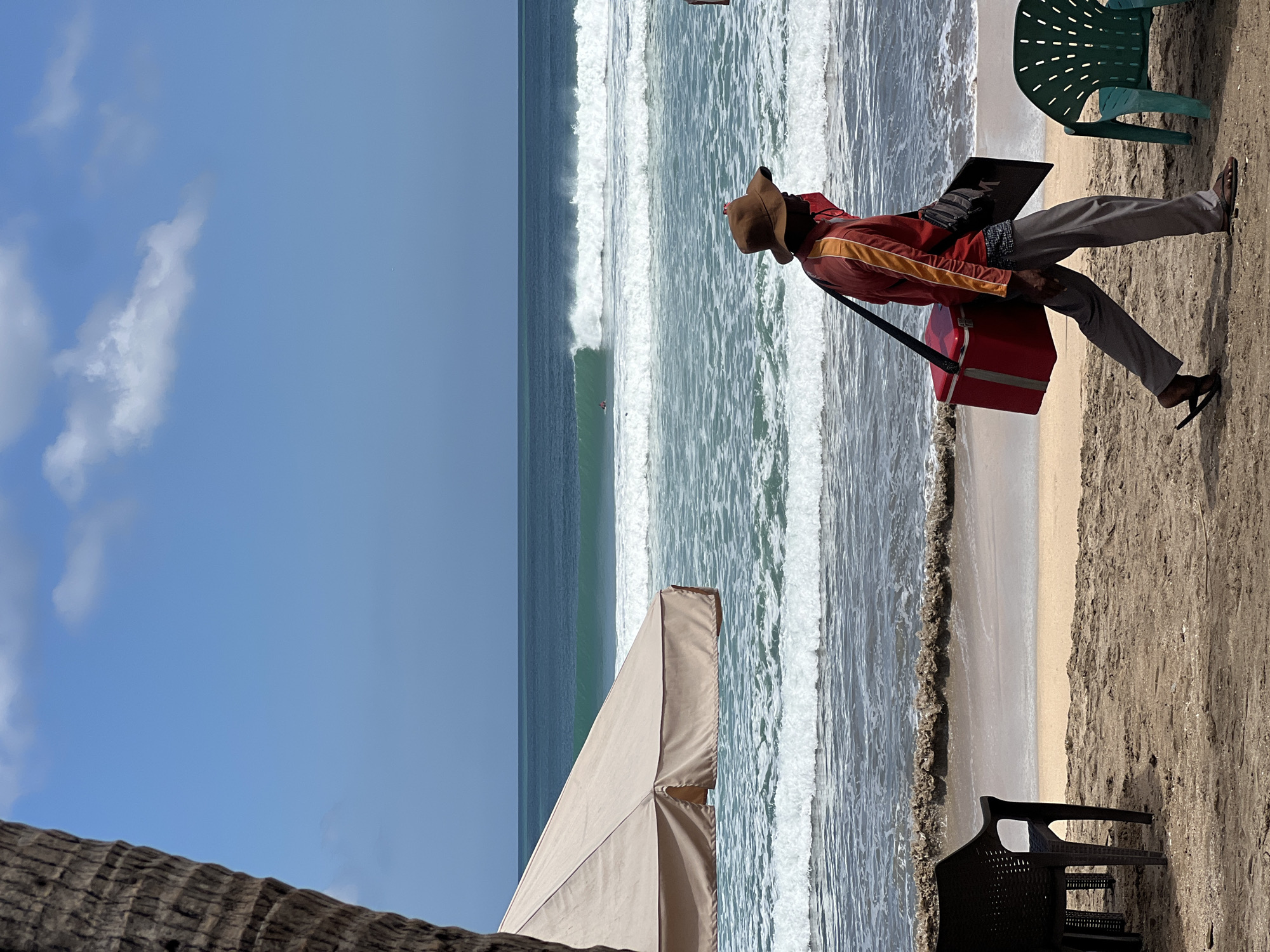 In Kuta, Indonesia, a person wearing a brown hat, an orange and red striped long-sleeved shirt, and gray pants walks across a sandy beach. They are carrying a red cooler bag slung over their left shoulder by a black strap, and a dark rectangular object is tucked under their right arm. The person is wearing dark flip-flops. In the foreground, a dark brown plastic chair is partially visible on the left, and a green plastic chair is on the right. In the background, the ocean has moderate waves breaking, with a light blue sky and some white clouds above. A beige beach umbrella is positioned to the left of the frame, casting a shadow on the sand.