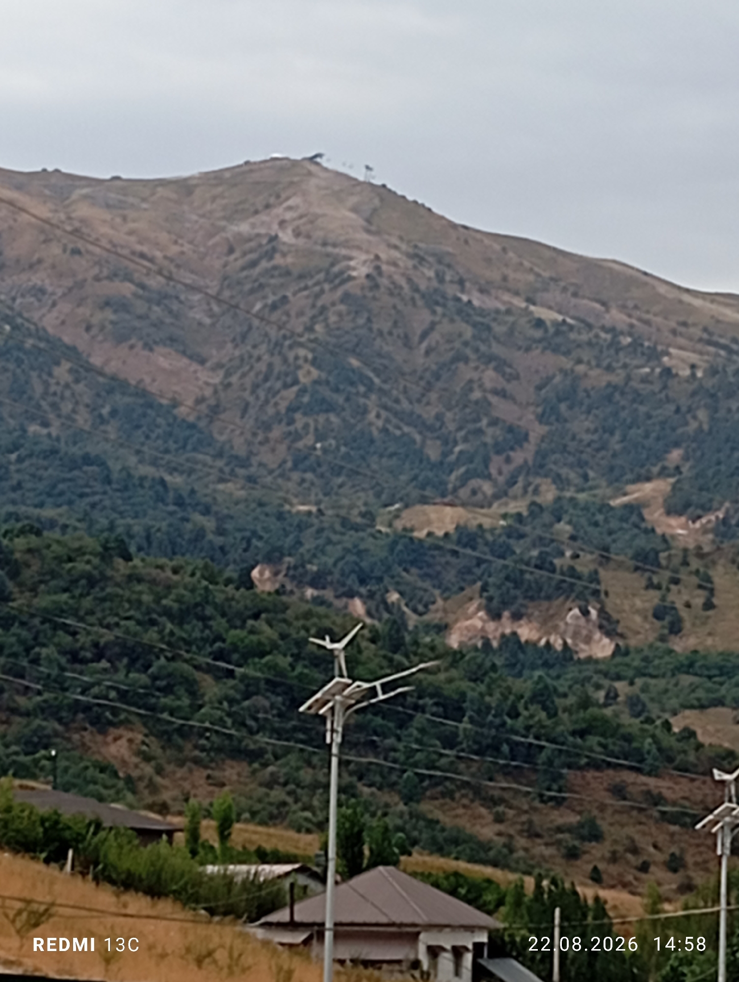 The image is a photograph taken in Amirsay, Uzbekistan. It depicts a mountainous landscape with trees and some buildings. The foreground shows a grassy area with a few structures, including what appears to be a solar-powered lamp post. In the mid-ground, there are houses and dense green trees covering the lower slopes of the mountains. The background is dominated by a large mountain with dry, rocky terrain and sparse vegetation. There are power lines running across the image. The sky is overcast. The timestamp "22.08.2026 14:58" and "REDMI 13C" are overlaid on the image. There are no people visible in the photograph.