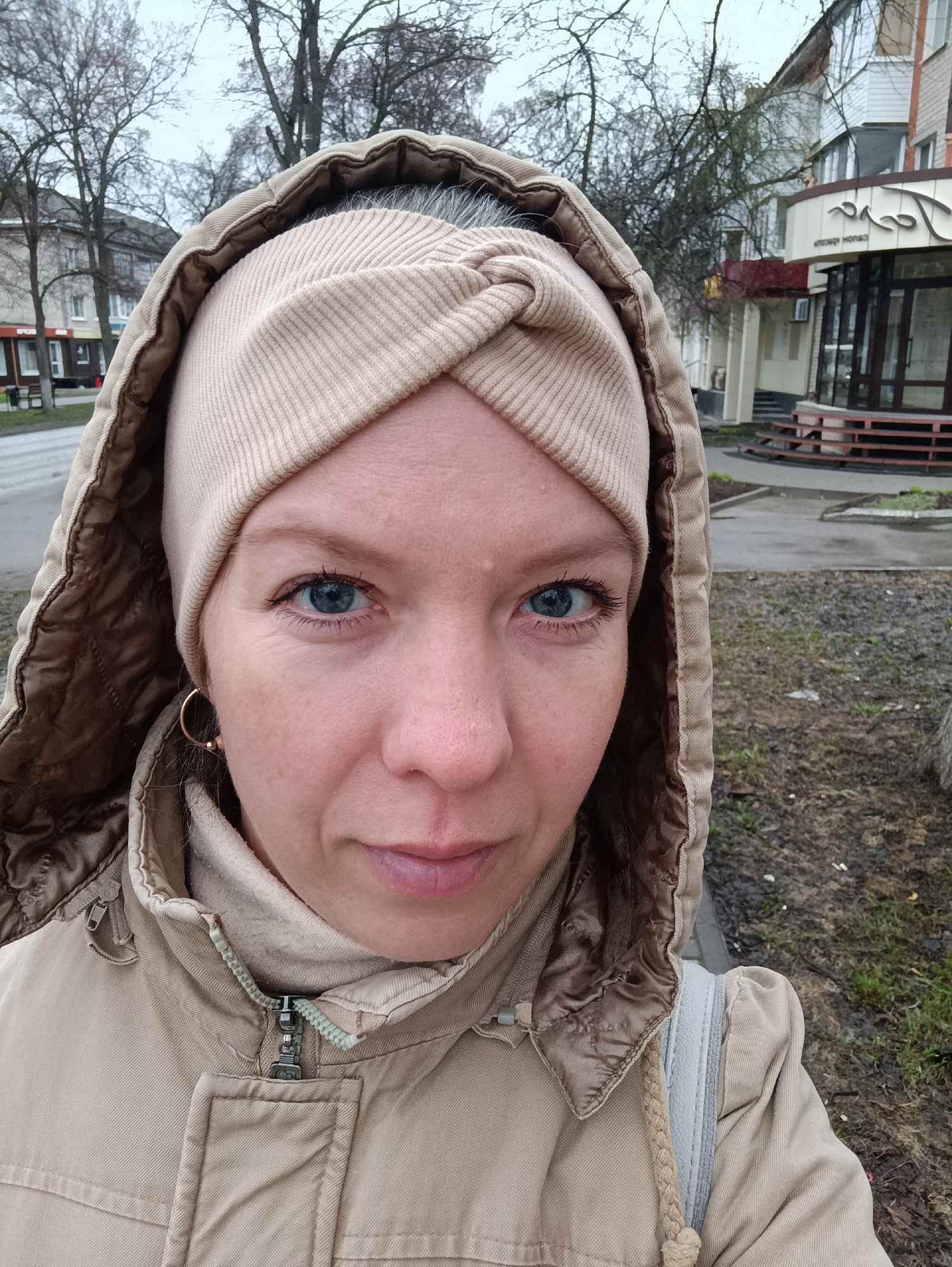 A woman is captured in a close-up, eye-level selfie outdoors in what appears to be Chaikovsky, Russia. She has light skin, blue eyes, and light-colored hair, some of which is visible under a light-colored, ribbed fabric headband. She is wearing a beige or tan hooded jacket with a contrasting brown lining inside the hood and a partially zipped front. Gold-colored hoop earrings are visible on her left ear. Her expression is neutral as she looks directly into the camera.
The background suggests an urban or suburban street scene on an overcast day. Bare trees, indicative of late autumn, winter, or early spring, are prominent behind her. To her left, buildings line a street, with one partially visible residential-style building. To her right, further back, another building with decorative trim features visible Cyrillic text above its entrance, which reads "САЛОН КРАСОТЫ" (Beauty Salon). The ground is a mix of bare soil, sparse grass, and paved areas, appearing damp. The overall lighting is soft and diffused, consistent with an overcast sky.