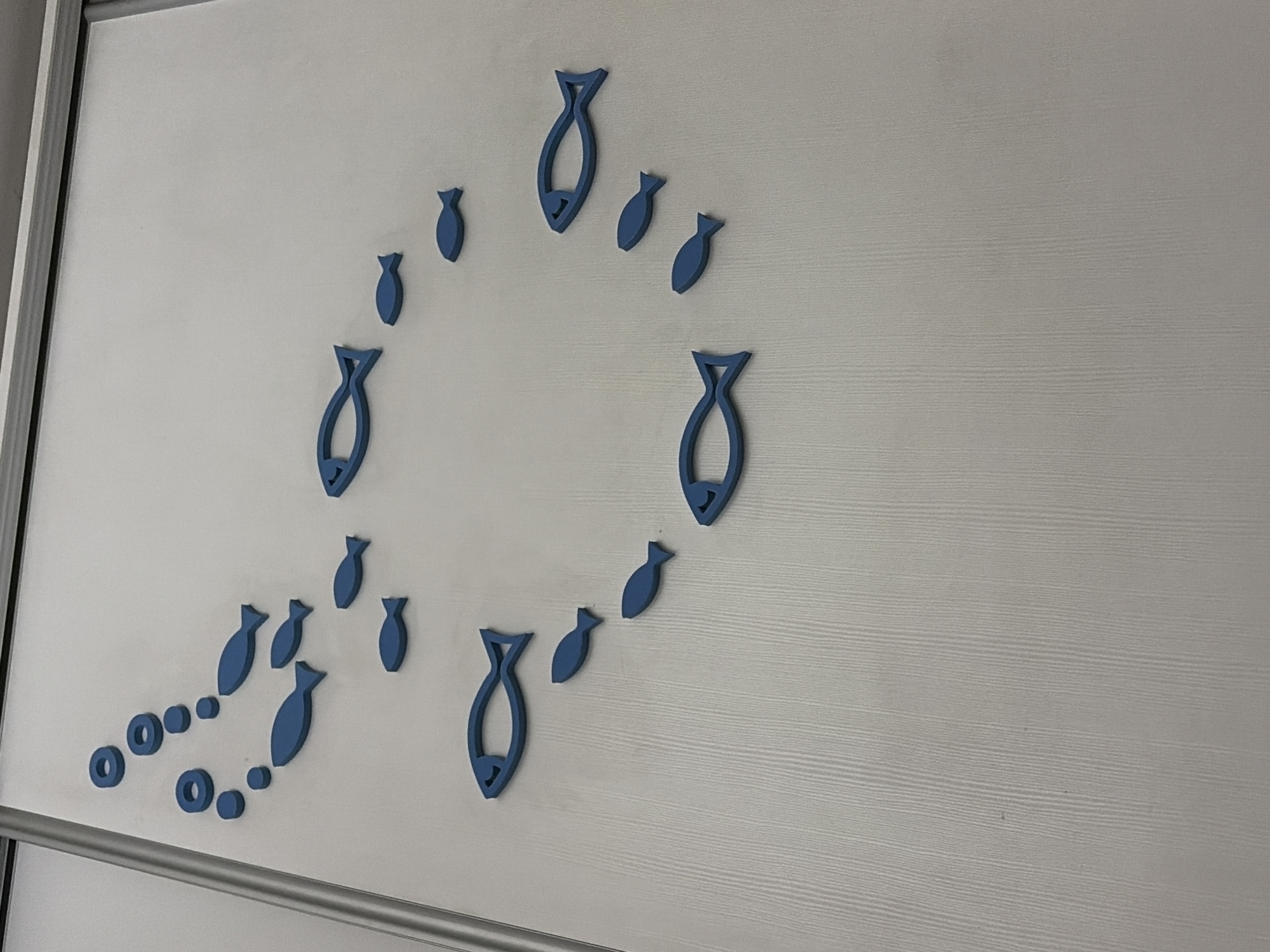 The image shows a close-up, eye-level view of blue, stylized fish cutouts and rings affixed to a textured white wall. The fish are arranged in a school-like formation, with some larger fish leading a trail of smaller ones. The lighting appears to be ambient, casting no strong shadows. The wall texture is a subtle, vertical wood grain pattern. No specific location in Vratsa, Bulgaria is identifiable from the image. The image is an indoor shot, likely on a wall in a room. There are no people or discernible actions taking place. The overall scene is static and decorative.