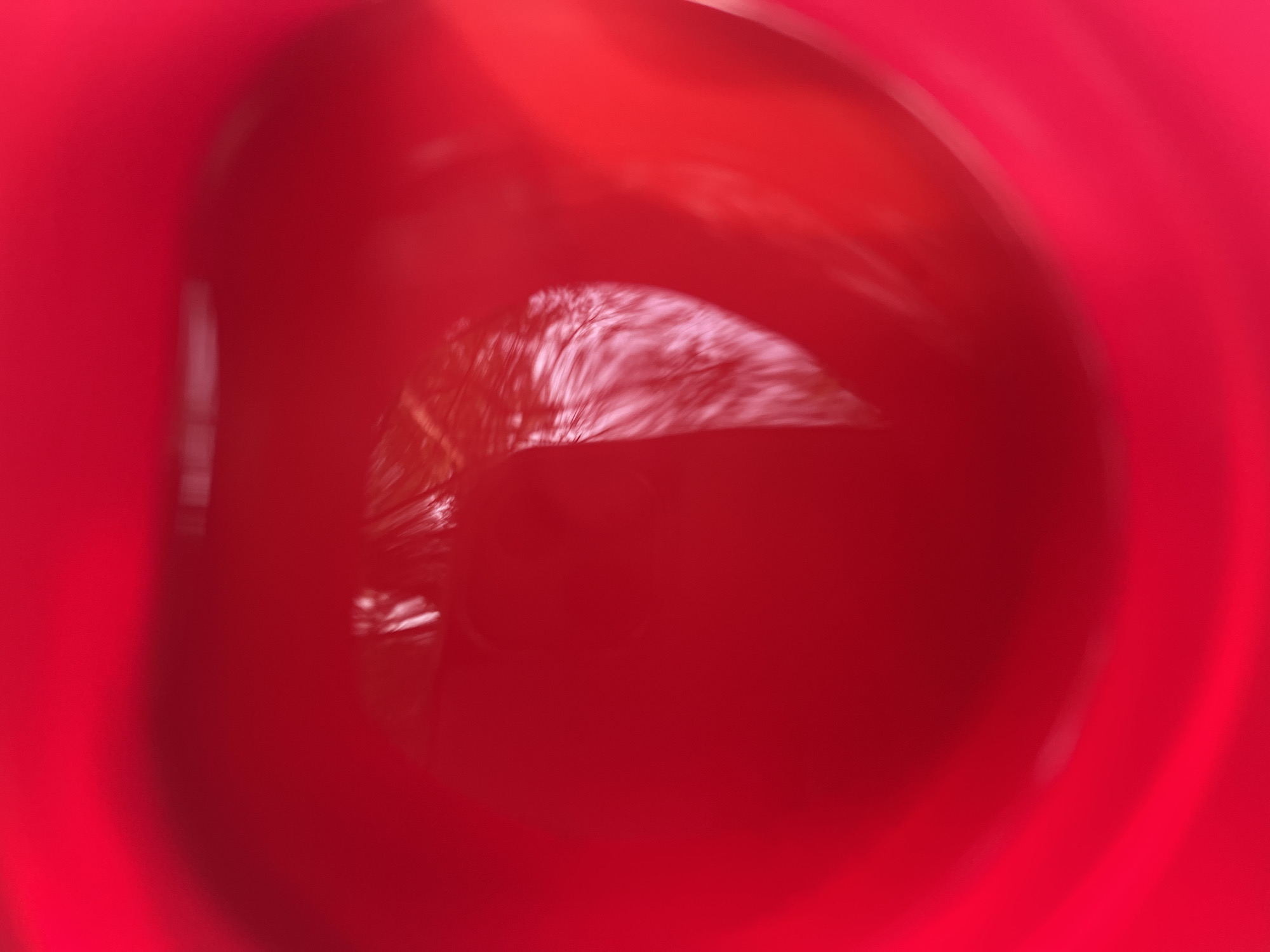 The image is a close-up, abstract shot of a red liquid, possibly wine or juice, in a glass or container. The liquid has a vibrant red color and reflects light, creating a glossy surface. The lighting is bright, causing some areas to appear blown out and others to be in shadow. There are subtle textures and ripples visible on the surface of the liquid, suggesting movement or the presence of fine particles. The overall impression is one of richness and depth, with a focus on the color and reflective qualities of the substance. There is no discernible location in the city of Raudoja, Estonia, or any contextual elements to place it there.