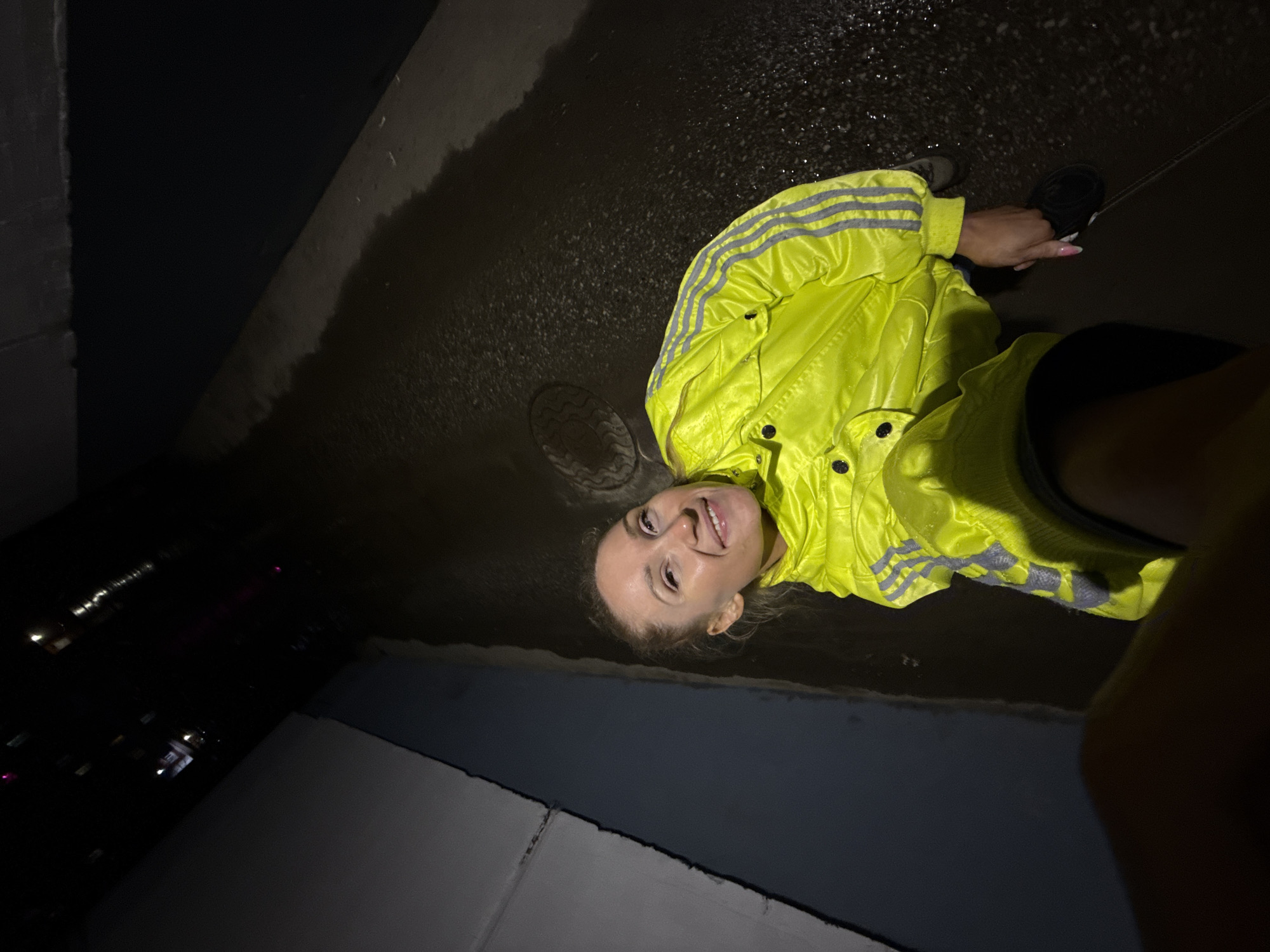A woman wearing a yellow jacket and black shoes is posing on a wet street at night. The background includes a sewer cover, a sidewalk, and building lights. The location is likely Perm, Russia.