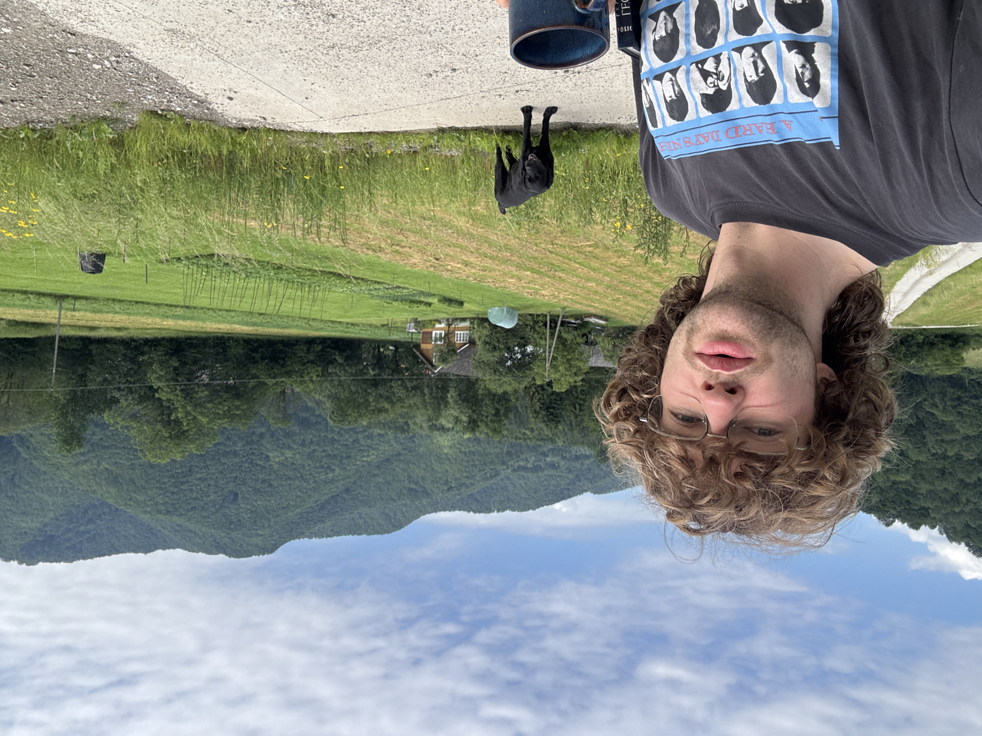 A young white male with curly brown hair and glasses is taking a selfie outdoors in a rural area. He is wearing a dark t-shirt with an image of The Beatles. To his right, a black dog stands on a concrete path, looking towards the camera. Behind them, a vast green landscape unfolds with rolling hills, a small house, a garden, and a road leading up a hill. The sky is partly cloudy.