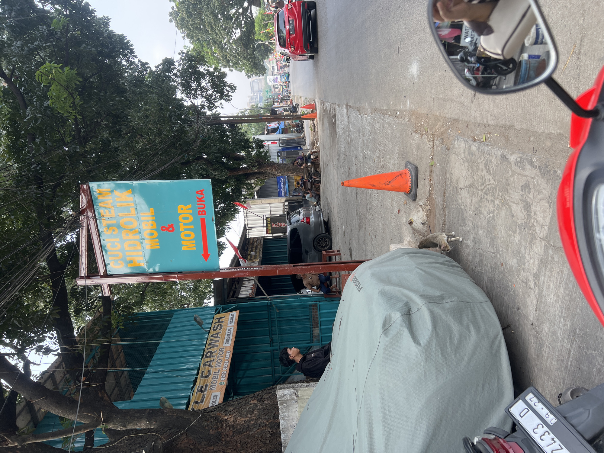 This is a street-level, eye-level, medium shot of a car wash and steam service located in Citeureup, Indonesia. The scene captures a bustling street environment with a prominent turquoise and orange sign advertising "CUCI STEAM HIDROLIK MOBIL MOTOR BUKA" (Steam Wash Hydraulic Cars Motorcycles Open).
On the left side of the image, a teal-colored building houses the car wash. A large tree with dense foliage partially obscures the building and a sign that reads "LE CARWASH MOBIL MOTOR" with a phone number and additional services. A young man wearing a black hoodie sits on a low wall to the left of the car wash entrance, looking towards the right. Next to him, a large gray car cover dominates the foreground. A cat is visible near the base of the car cover, positioned between it and the sidewalk.
The right side of the image shows a paved road with traffic cones and a red car driving away from the camera. Several people are seated on a bench in front of a building on the right side of the road, appearing to be patrons or workers at a nearby establishment. Beyond them, a street stretches into the distance, lined with various businesses and trees. The sky is overcast, suggesting a cloudy day. The overall impression is a typical street scene in a developing urban area, with a focus on local businesses and everyday life.