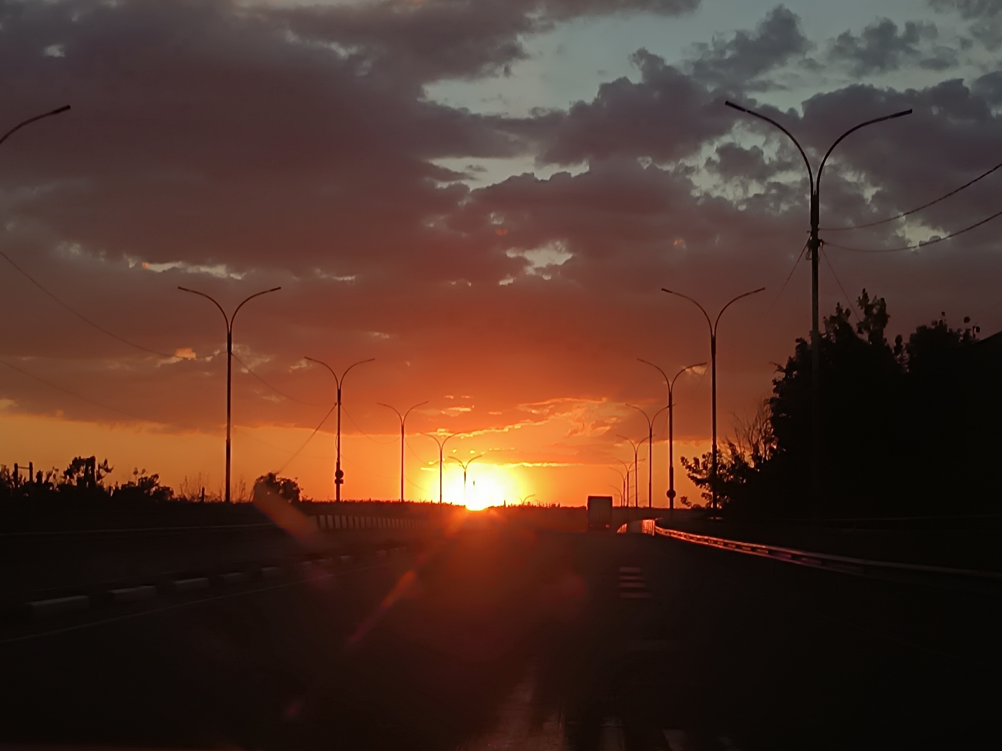 The image depicts a highway at sunset. The sun is low on the horizon, casting a warm orange glow across the sky and reflecting off the wet road surface. Numerous streetlights line both sides of the highway, their silhouettes stark against the vibrant sky. The clouds are a mix of dark gray and fiery orange, adding to the dramatic atmosphere of the scene. The road appears to be clear of traffic, with only a single truck visible in the distance. The overall impression is one of a serene and beautiful end to the day, observed from the perspective of a moving vehicle.
