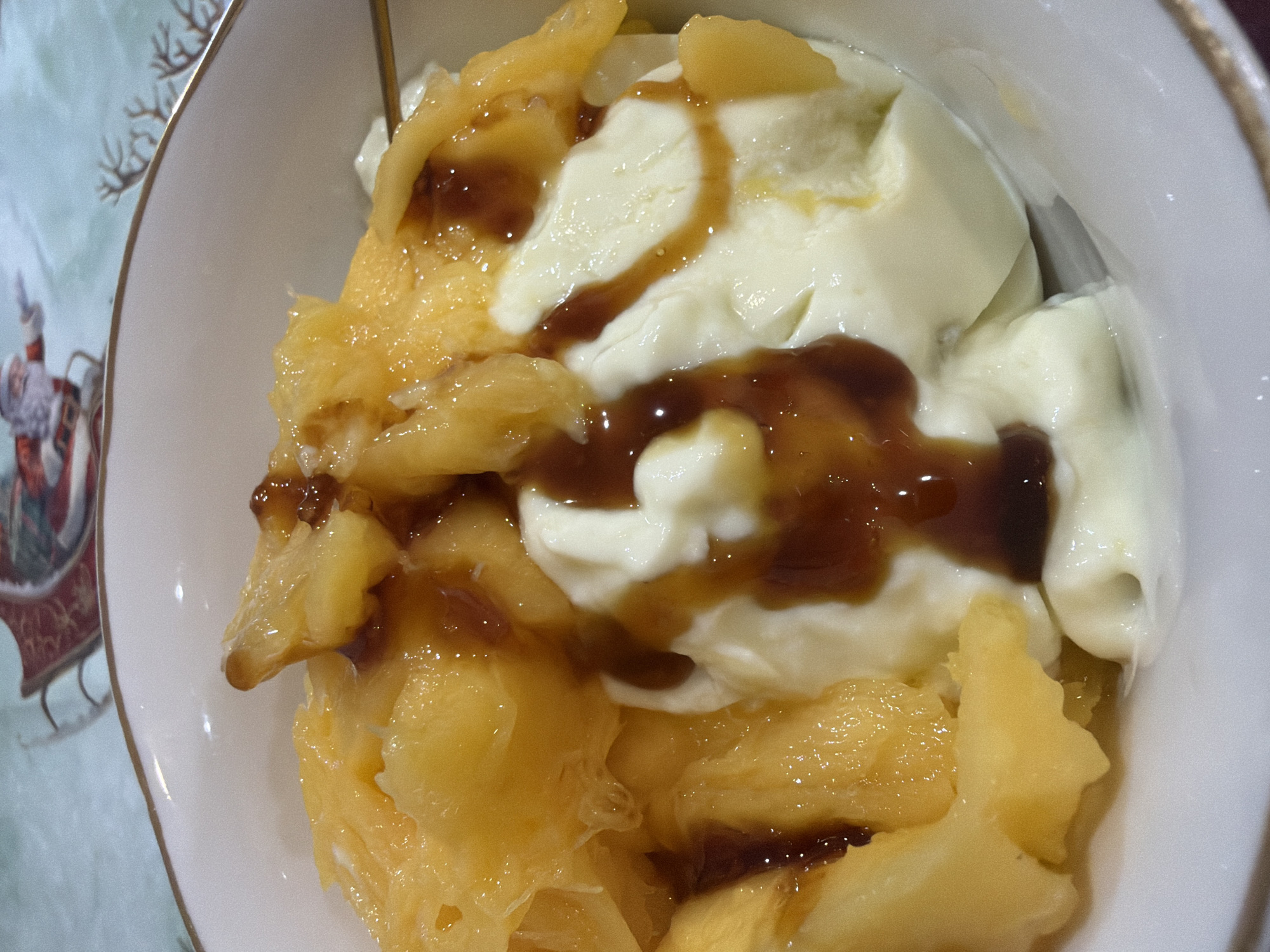 A close-up shot of a dessert in a white bowl. The dessert consists of diced yellow fruit, possibly pineapple or mango, topped with scoops of white, creamy substance like yogurt or ice cream and drizzled with a dark brown sauce, likely caramel or chocolate. The bowl has a decorative gold rim. The edge of a tablecloth or placemat with a Christmas-themed design is visible in the background. The picture's location is not discernible from the visual information provided.