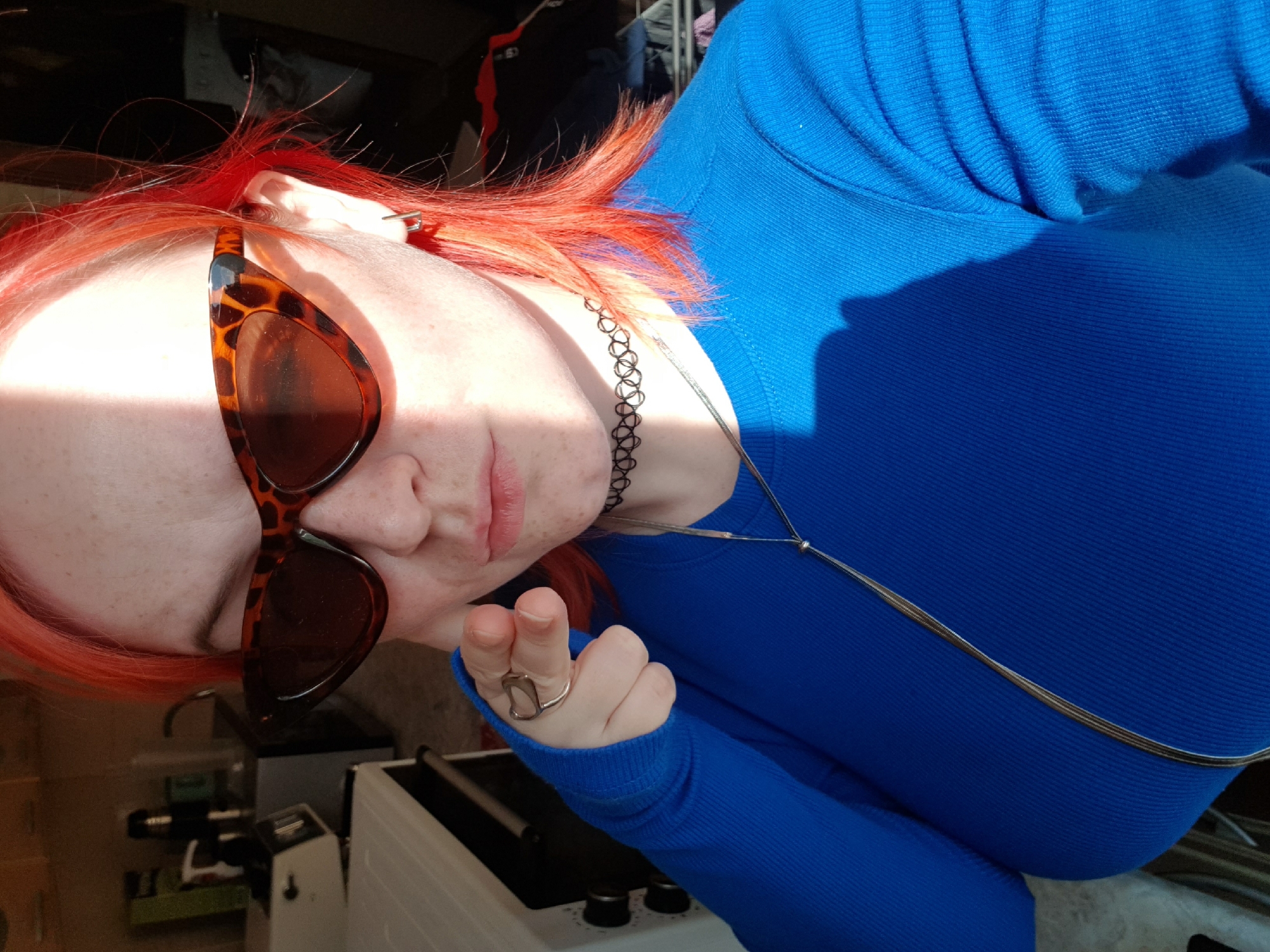 A person with red hair is wearing sunglasses and a blue shirt. The location is Unknown, Unknown. The person is wearing a necklace and a choker. There is a ring on their finger. A white machine with knobs is visible in the background. The person is looking down at the camera.