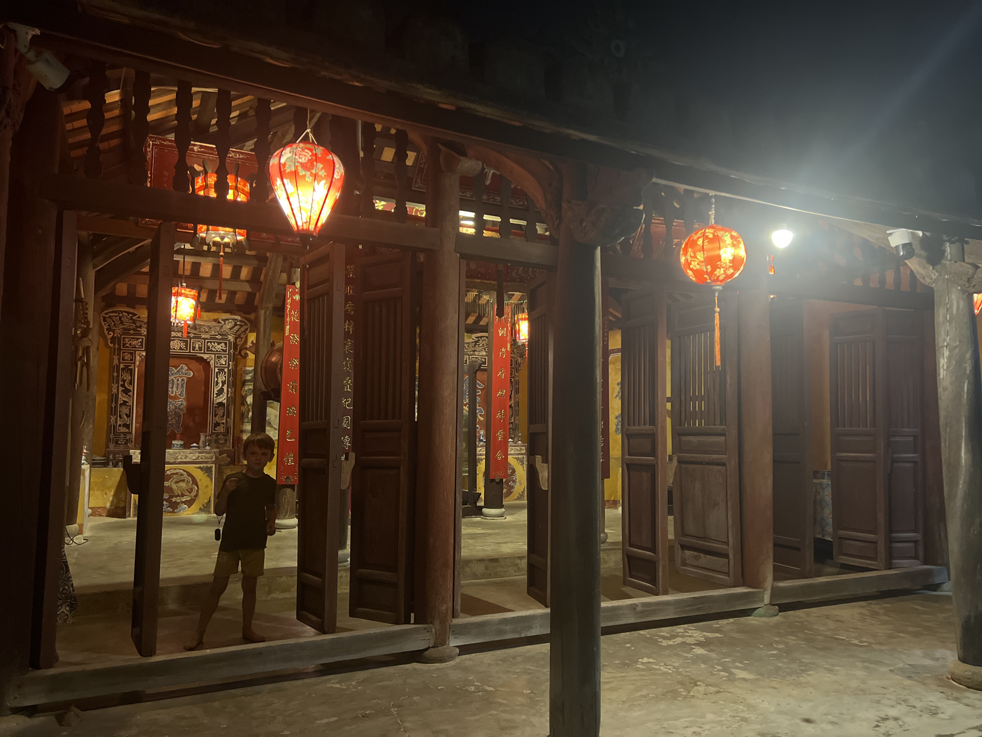 A young boy stands in the doorway of an ancient building in Hoi An, Vietnam, illuminated by traditional red lanterns. The boy is wearing a dark t-shirt and light-colored shorts, with his left hand held up to his face. He is barefoot. The building features dark wooden pillars and walls, with open doorways revealing an interior adorned with ornate decorations and more lanterns. The ground is paved with concrete, and the scene is lit by the warm glow of the lanterns, creating a mystical atmosphere. The architecture suggests a temple or ancestral house, typical of Hoi An's historic Old Town.