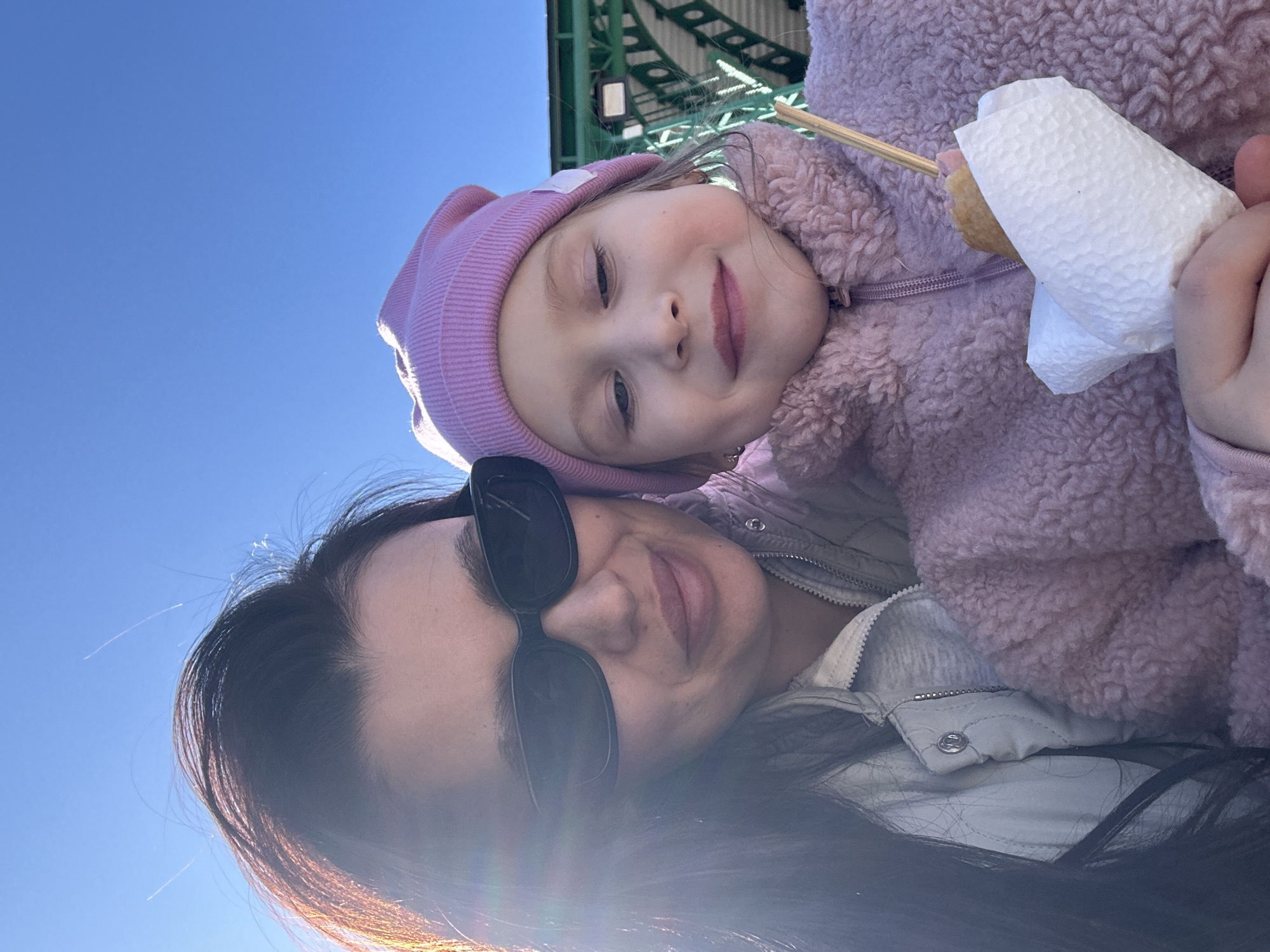 A selfie-style image depicts a woman and a young girl. The girl, wearing a pink hat and a fuzzy pink jacket, holds a food item wrapped in a white napkin on a stick, suggesting a corn dog or similar snack. The woman wears sunglasses and her dark hair is visible. The background features a clear blue sky and architectural elements that look like a bridge or structure with green beams and white detailing. The location is the city of Chaikovsky, Russia. The photograph appears to have been taken outdoors on a sunny day. The primary action depicted is the taking of a selfie with a child.