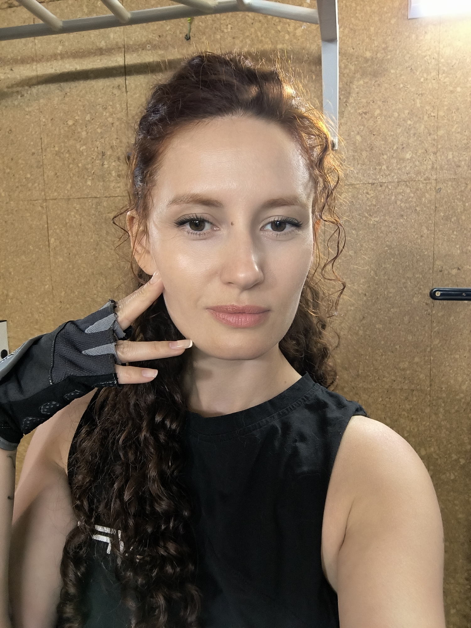 A woman with curly, reddish-brown hair and light skin is posing for a selfie in what appears to be a gym. She is wearing a black sleeveless top and a black workout glove on her left hand. Her left hand is touching her cheek. The background is a corkboard wall with a metal bar overhead and a black object mounted on the wall to the right. The lighting suggests it is daytime, possibly indoors. Her expression is neutral and she is looking directly at the camera. There is a subtle mole on her right cheek. The woman has defined eyebrows and is wearing light makeup. The overall impression is a candid, post-workout moment.