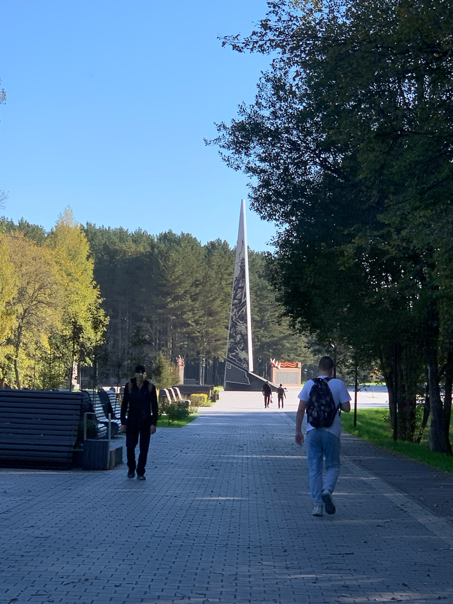 This is a full-frame, eye-level shot of a paved walkway in a park setting. In the foreground, two men are walking away from the camera. The man on the left is wearing a black jacket and dark pants. The man on the right is wearing a white t-shirt, light blue jeans, and a backpack with a camouflage pattern. The walkway is made of light gray bricks and is lined with dark wooden benches on the left.
In the midground, there is a tall, dark, angular monument or sculpture with a white tip that tapers to a point. The monument features a detailed, light-colored relief or mural on its surface. Behind the monument, a line of dense green trees forms a backdrop. To the left of the monument, a smaller structure with red accents is visible.
The background consists of a clear blue sky and a dense forest of tall, green pine trees. The lighting suggests it is daytime, likely late morning or early afternoon, with shadows cast by the trees and walkway. The park appears to be a public space, possibly a memorial park given the monument. Based on the information provided, this scene is located in the city of Prokopyevsk, Russia.