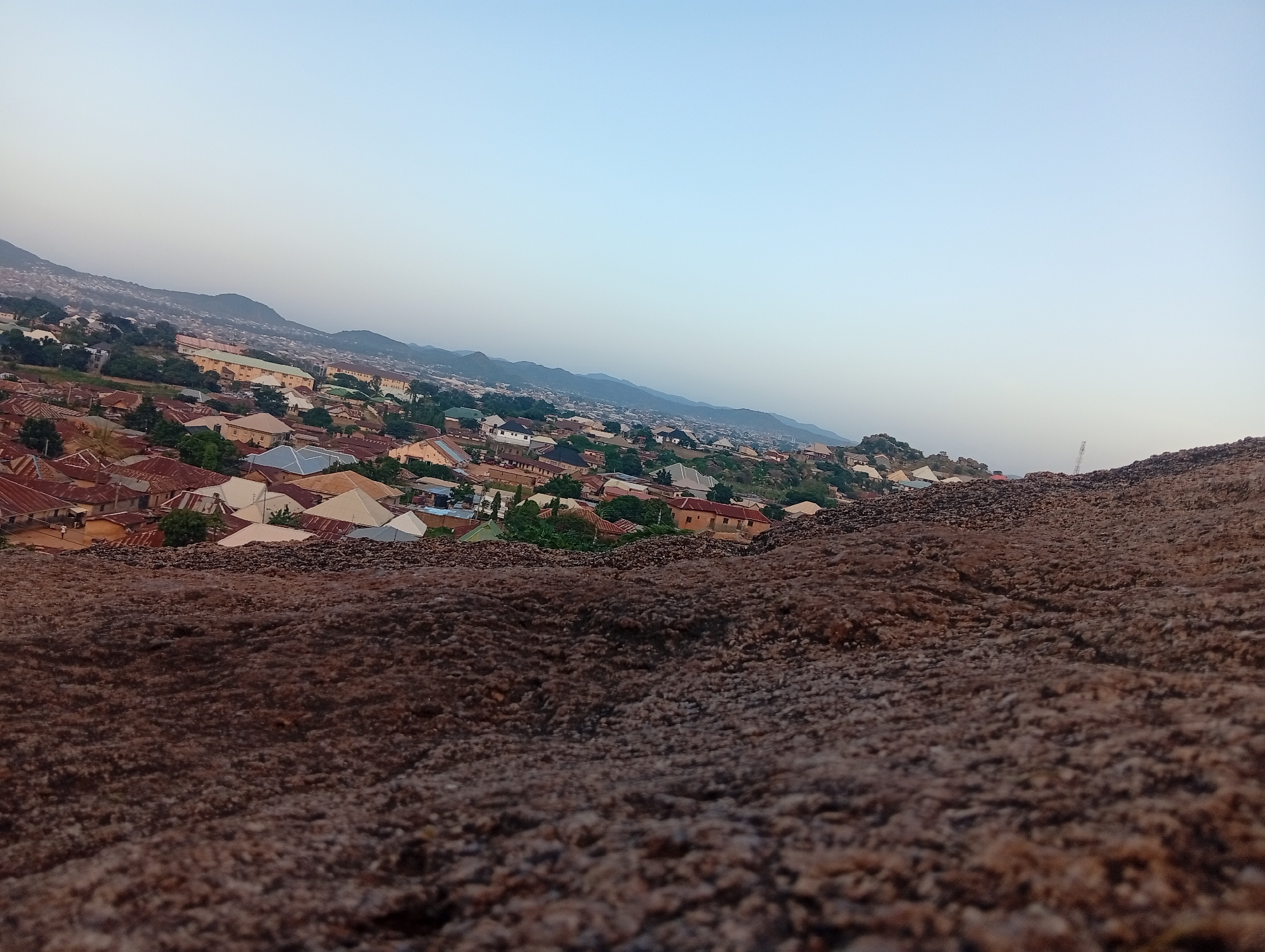 The image depicts an aerial view of a densely populated urban area, likely in the city of Kabong, Nigeria, as suggested by the context. The foreground is dominated by a rough, rocky surface, suggesting a natural elevation or viewpoint. Beyond this, the landscape unfolds into a sprawling cityscape characterized by numerous residential buildings with distinct red and white corrugated iron roofs. The architecture appears to be a mix of traditional and modern styles, with some structures featuring flat roofs and others with peaked designs.
Green vegetation is interspersed throughout the urban environment, indicating parks, gardens, or natural growth within the city. In the distance, a range of hills or mountains forms the backdrop, their slopes also appearing to be populated with houses, creating a sense of the city extending into the surrounding terrain. The sky above is a pale, clear blue, suggesting it is either early morning or late afternoon, with the light casting soft shadows. No people are clearly visible in the image, and the scene is static, capturing a moment of quiet observation over the city.