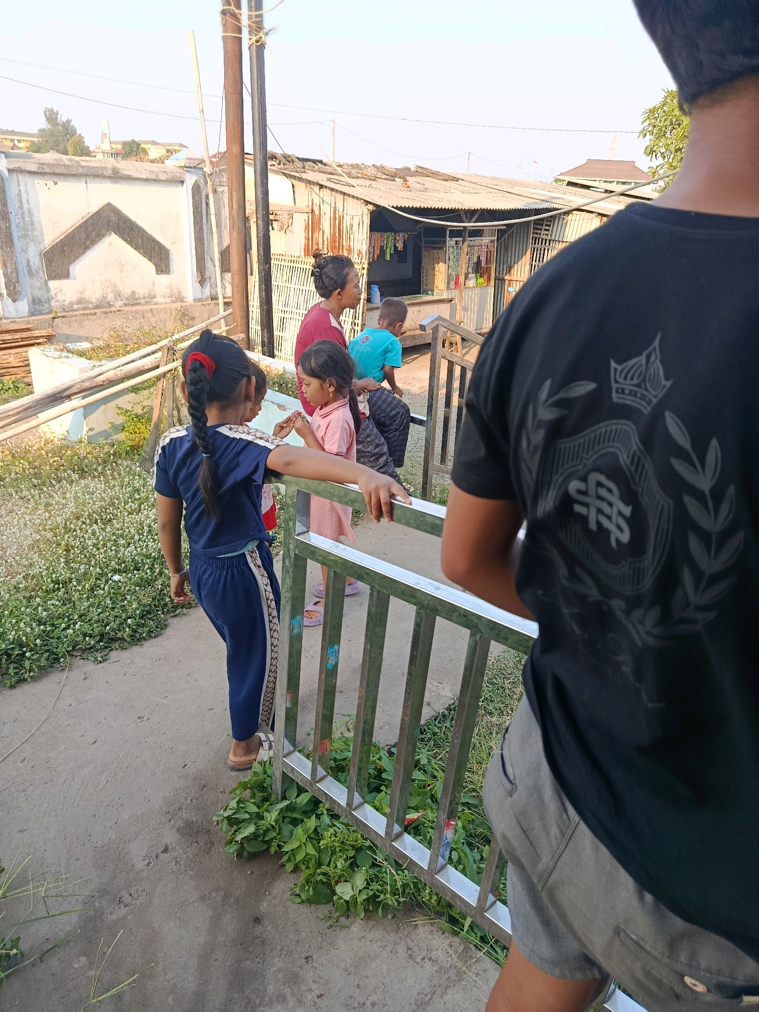 In Jobokuto, Indonesia, on a sunlit day, a young girl in a blue tracksuit with a red hair tie stands facing away from the camera, leaning on a metal railing. She has a dark braid down her back. Beside her, a younger child in a pink shirt and dark pants is interacting with another child in a turquoise shirt. Behind them, an adult, likely female, with her hair in a bun, wearing a reddish-pink shirt, looks on. In the background, a rural or semi-urban setting is visible with modest buildings, some appearing to be made of corrugated metal and wood, possibly shops or dwellings. The environment includes a concrete pathway, a patch of green ground cover with small white flowers, and various construction materials. A large wooden pole with wires runs vertically on the left side of the frame. The focus of the image is on the group of children and the adults, suggesting a moment of everyday life or transit.