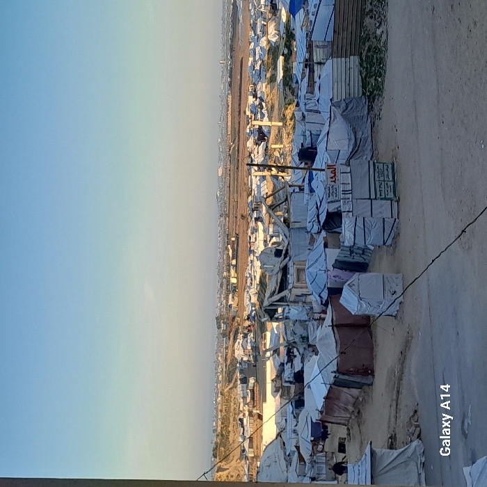 A crowded refugee camp extends to the horizon under a clear sky. Makeshift shelters, tents, and temporary structures are densely packed. A dirt road runs along the side of the camp. Boxes and supplies are stacked, some labeled with Arabic script. The landscape suggests a dire humanitarian situation.
