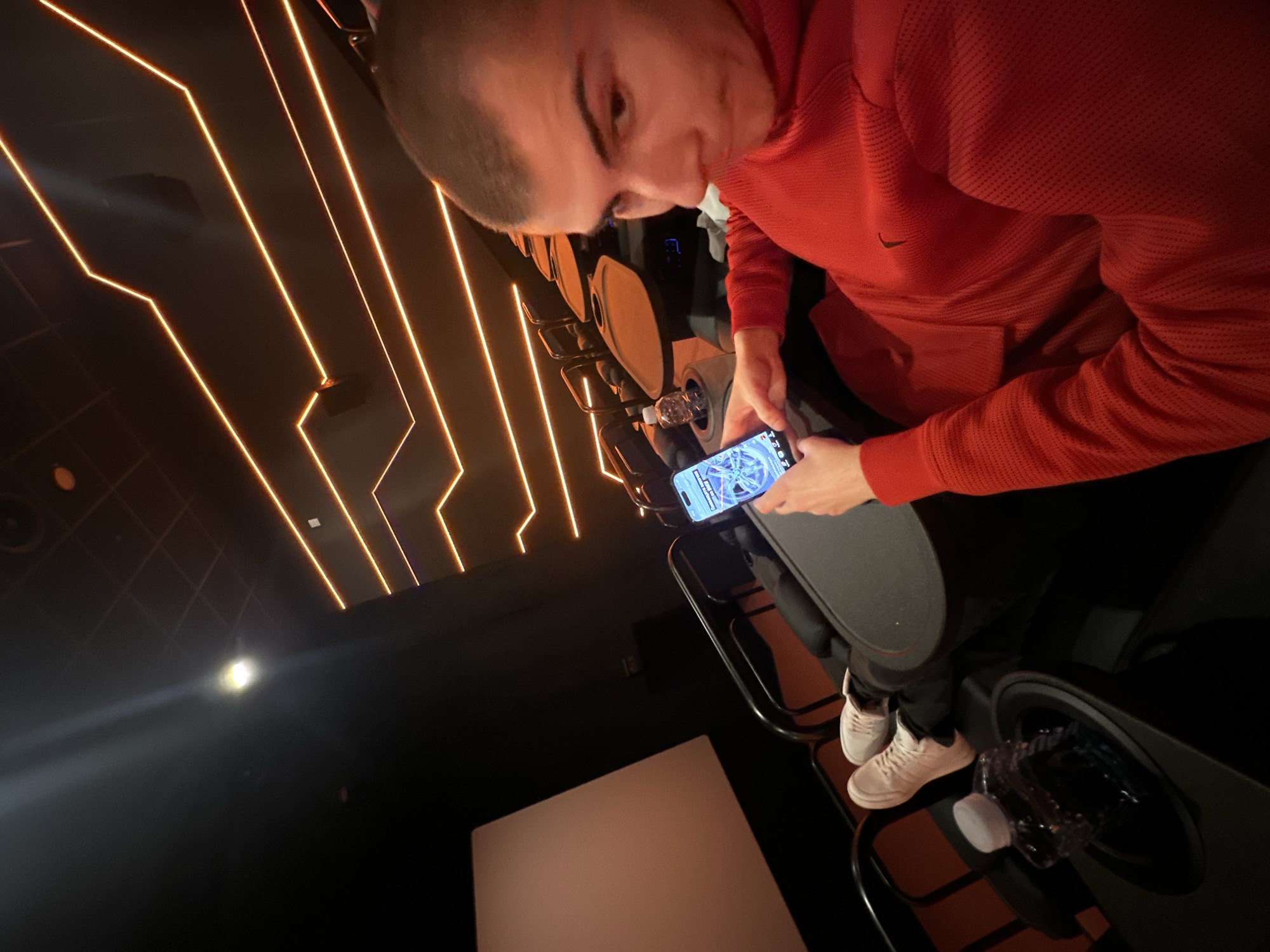 A person is seated in a dark environment that appears to be a movie theater or similar venue with tiered seating. The person is holding a mobile phone and looking at the screen. They are wearing a red hooded shirt and white sneakers. There is a water bottle in a cupholder nearby. The walls are dark with geometric orange lighting fixtures. The specific location in Plovdiv, Bulgaria, cannot be determined from the image.