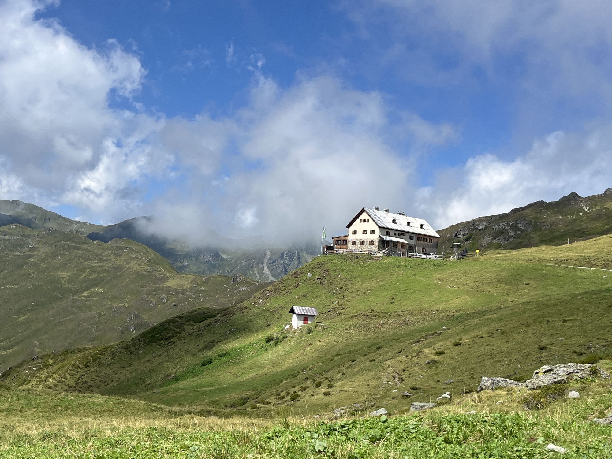 The image depicts a panoramic view of a mountain landscape in Hippach, Austria. In the mid-ground, a large, multi-story building with a white roof and light-colored walls sits atop a lush green hill. The building appears to be a mountain hut or lodge, with visible windows and balconies. In front of the lodge, a smaller, red-roofed structure with white walls is visible. The surrounding environment consists of rolling, verdant hills and rugged mountain peaks in the background, partially shrouded by clouds. The sky is a vibrant blue with scattered white clouds. The foreground is dominated by a grassy meadow with rocks scattered throughout. There are no people visible in the image.