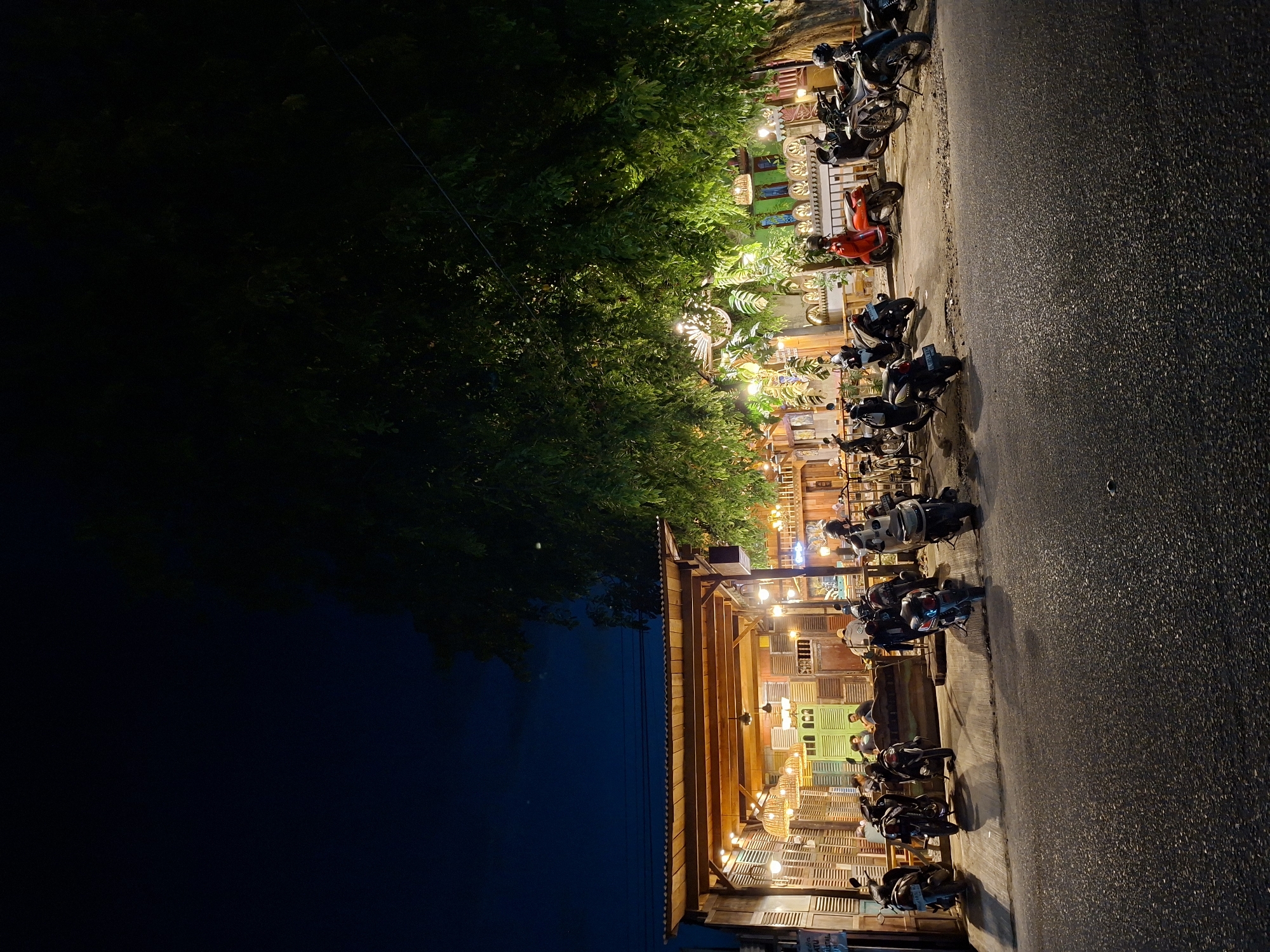 At night in Lhokseumawe, Indonesia, a row of parked motorcycles line the edge of a road next to a brightly lit open-air building. The building's façade is constructed from repurposed wooden shutters of various colors. Overhead string lights illuminate the building's exterior and the adjacent sidewalk. Lush green trees extend above the building, partially obscuring the night sky. The road surface appears dark and textured.