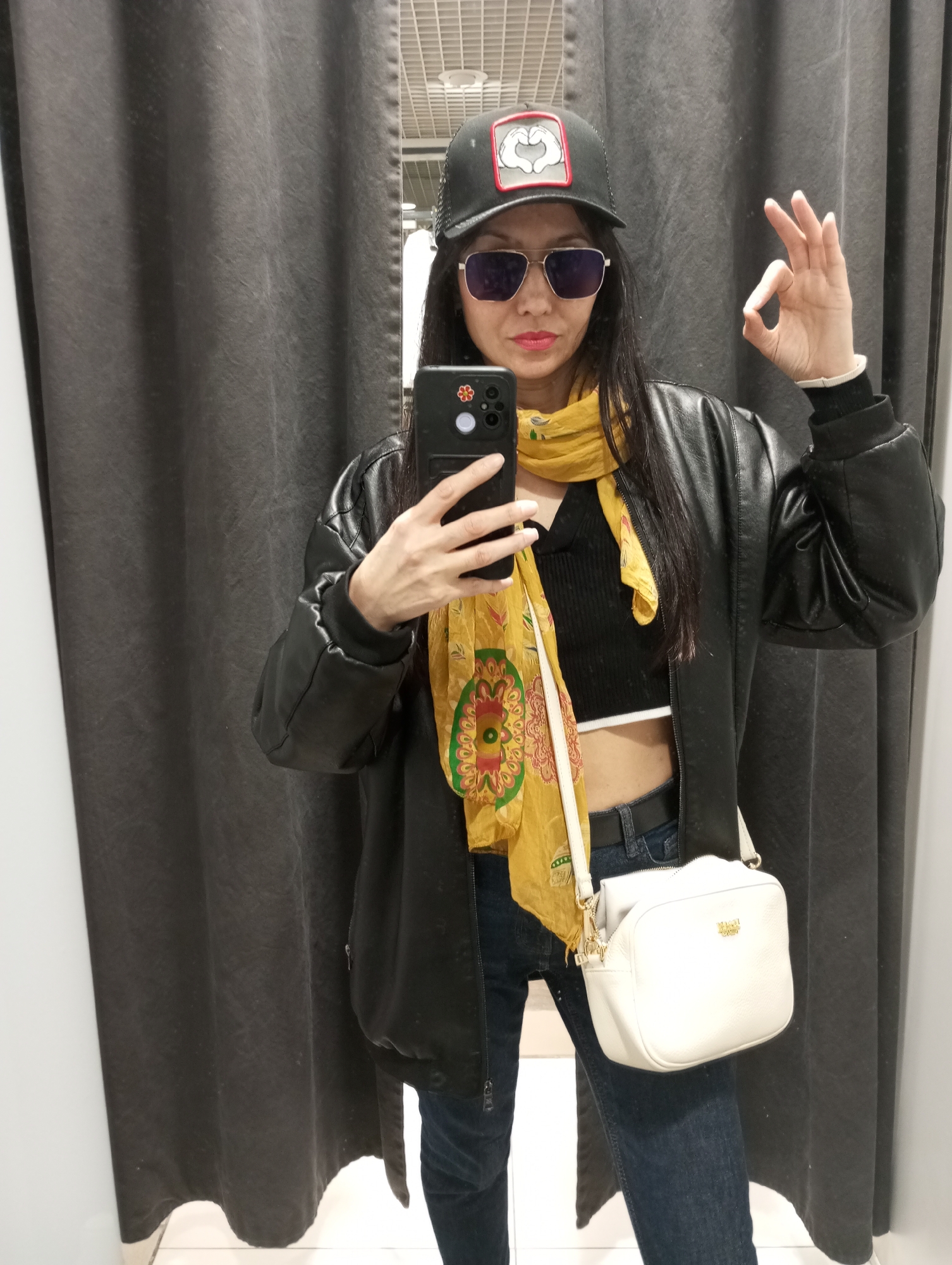 A woman is taking a selfie in a dressing room. She is wearing a black baseball cap with a logo on the front, aviator sunglasses, a black leather jacket, a black crop top, blue jeans, and a yellow scarf. She has a white purse slung over her shoulder. She is holding her phone in her left hand and making an "OK" sign with her right hand. The dressing room has a grey curtain and a white floor. It is unknown if the location is in Izhevsk, Russia.