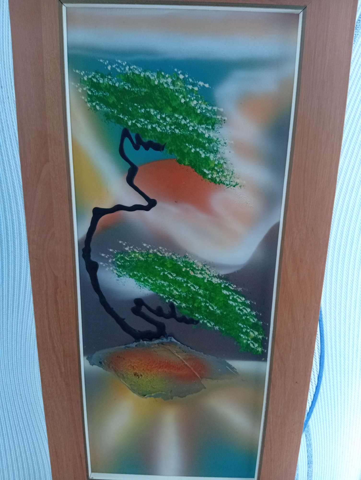 A framed spray-painted artwork is shown vertically, with a wooden frame surrounding a colorful abstract scene. The artwork depicts a stylized tree with a dark, winding trunk and green foliage dotted with white specks. The tree appears to be growing from a rock-like formation at the bottom, which is rendered in shades of orange, yellow, and red. The background is a blend of abstract colors, including blues, oranges, and grays, creating a sense of depth and atmosphere. The artwork is positioned against a light blue and white striped background, possibly a wall or fabric. The overall impression is a whimsical and artistic depiction of nature.