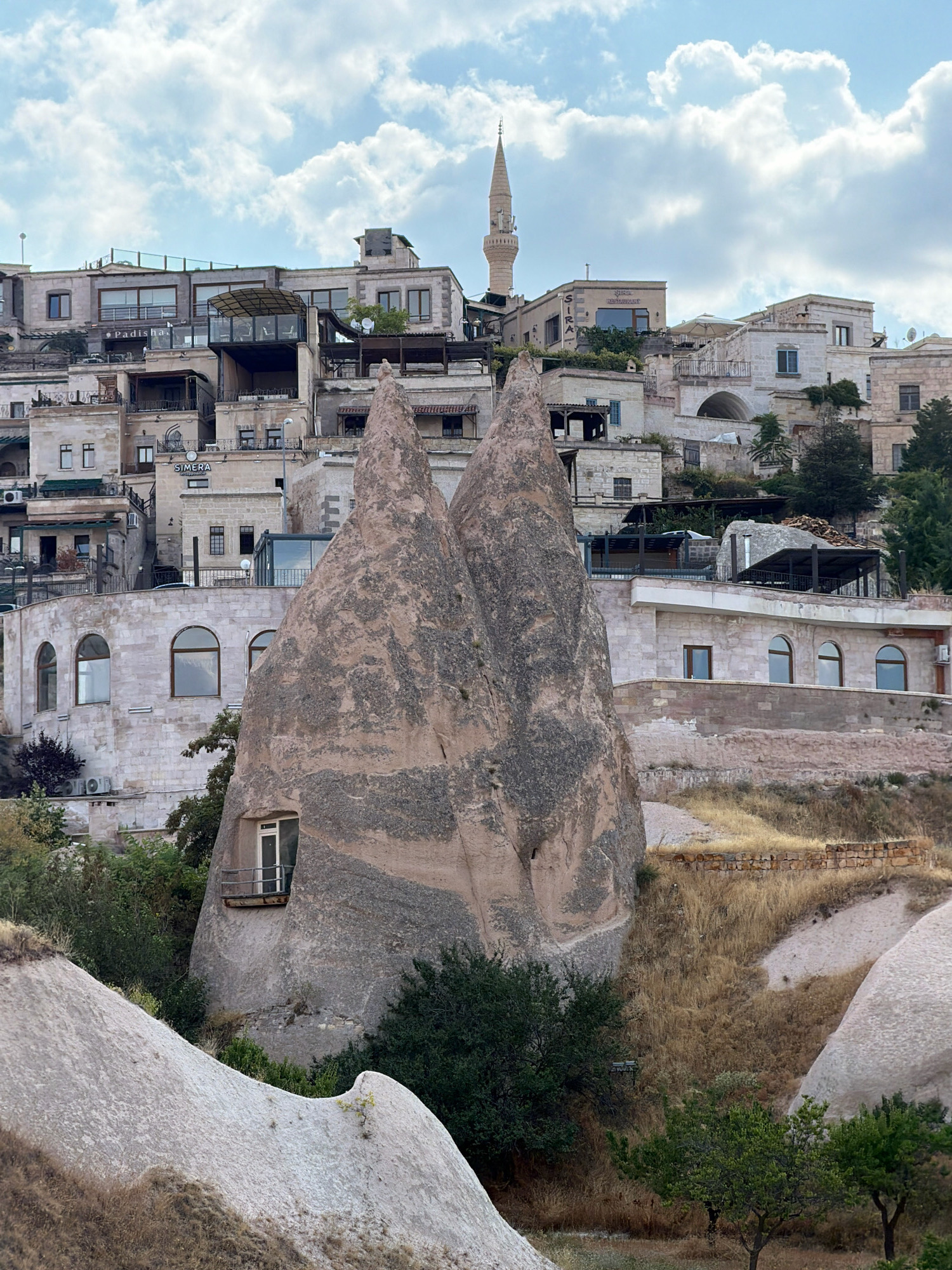 This image captures a view of the unique landscape of Uçhisar Beldesi, Turkey, characterized by its distinctive rock formations and ancient architecture. The foreground features several of these iconic "fairy chimneys," with their weathered, cone-like shapes. One prominent chimney in the center has a small balcony and a window carved into its side, suggesting human habitation within the rock.
Behind the rock formations, the town of Uçhisar is visible, built into and around the natural landscape. Buildings are constructed with light-colored stone, featuring multiple levels, terraces, and arched windows. Some structures appear to be modern hotels or guesthouses, indicated by balconies, awnings, and signage. A minaret, a common architectural feature in Turkey, rises from the cluster of buildings, with its slender tower and pointed cap reaching towards the sky. The sky is partly cloudy, with large, white cumulus clouds against a pale blue background. The terrain is dry and arid, with patches of grass and some scattered trees and bushes adding touches of green to the earthy tones of the rocks and buildings. The overall impression is one of a place where human civilization has adapted to and integrated with a dramatic and ancient natural environment.
