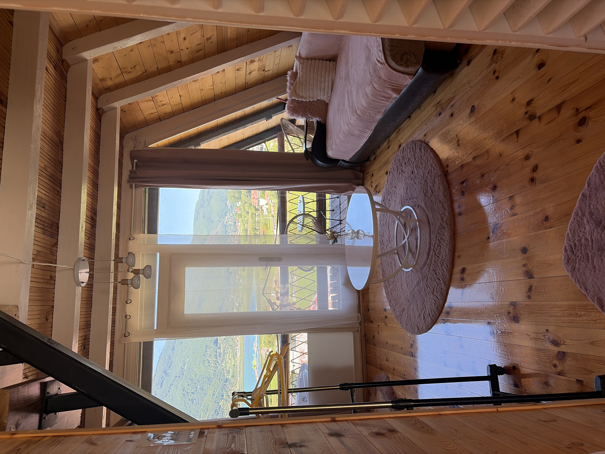 This is a high-angle, interior shot of a room with a view of Plav Lake in Plav, Montenegro. The room features a wooden sloped ceiling and walls, a polished wooden floor, and a large window overlooking the lake and surrounding mountains. In the foreground, a black garment rack with yellow hangers stands to the left. A circular pink rug is in the center, with a white circular coffee table on top. To the right, a pink sofa is adorned with two throw pillows, one pink and fluffy, the other cream-colored and ribbed. A sheer white curtain hangs in front of the window, partially obscuring the view. Outside the window, balconies and chairs are visible, along with the picturesque landscape of Plav Lake. The overall impression is one of a cozy, rustic accommodation with a stunning natural backdrop.