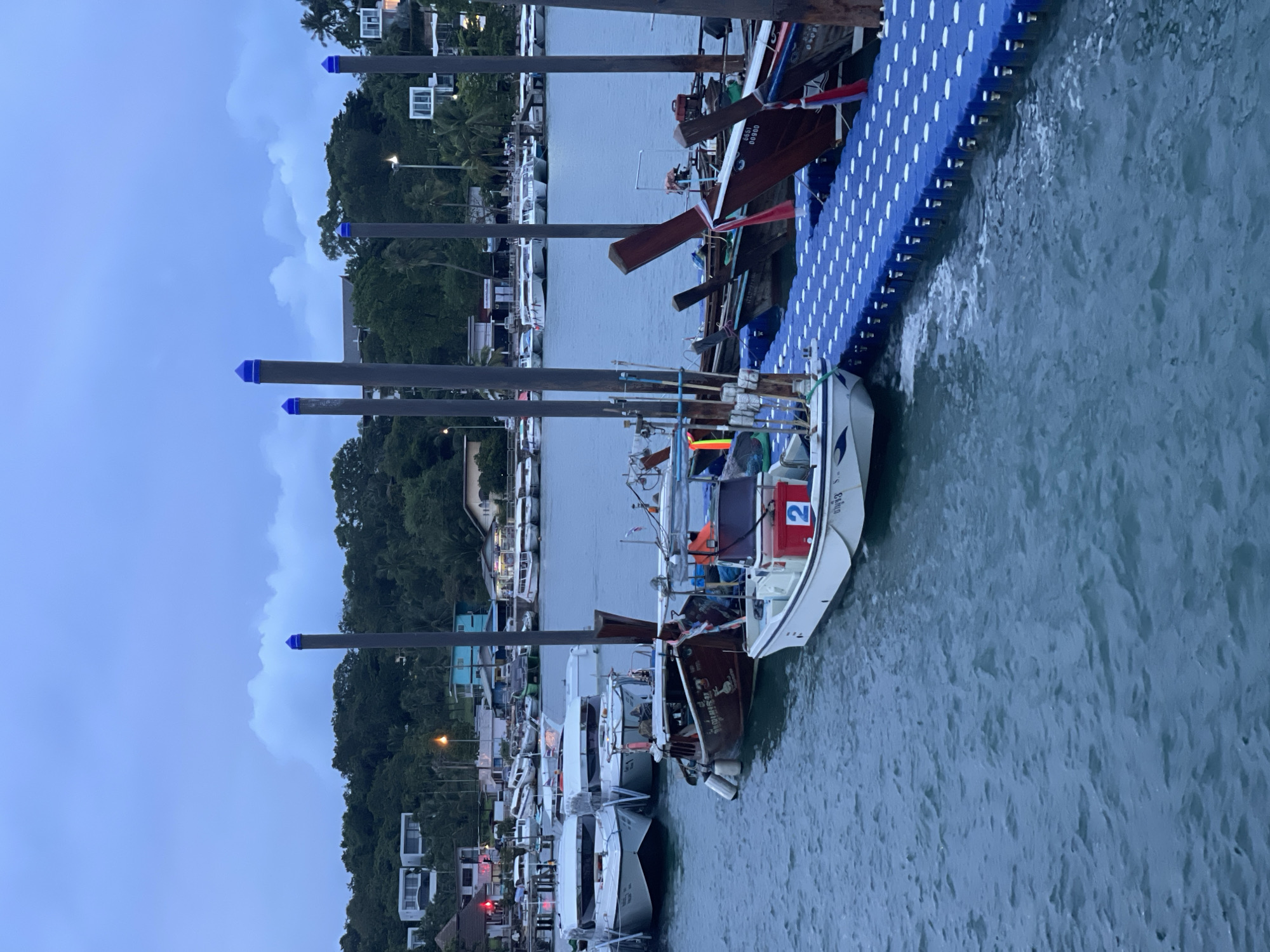 The image shows a row of docked boats on a body of water, likely a canal or harbor, in Pa Khlok, Thailand. The scene is set at dusk or dawn, as indicated by the dim, overcast sky and the artificial lighting visible in the background. Several boats are lined up along a blue, modular floating dock. The boats vary in size and style, with some appearing to be fishing vessels and others more recreational. One boat in the foreground has the number "2" prominently displayed on a red box. Another boat further back has text written on its hull. The surrounding area features lush green trees and vegetation covering a hilly landscape, with buildings and houses scattered along the shoreline. The water is calm with gentle ripples. The overall atmosphere is serene, with a sense of quiet activity associated with a harbor at the beginning or end of the day.