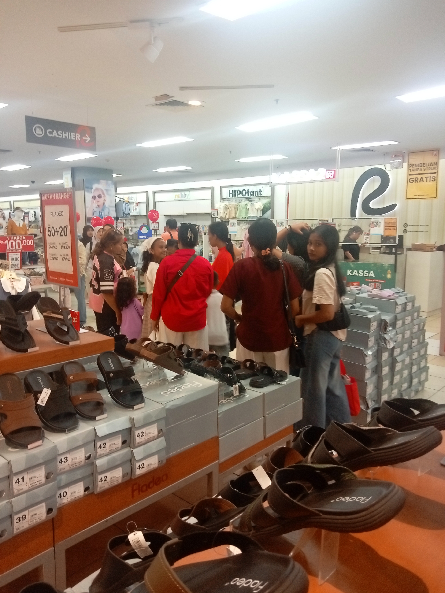 The image depicts an indoor shoe store, likely in Batam, Indonesia, displaying various styles of footwear. In the foreground, multiple pairs of sandals are arranged on shelves, some presented on clear acrylic stands. Cardboard boxes, labeled with shoe sizes and the brand "Fladeo," are stacked neatly behind the displayed sandals. Further into the store, a group of people are browsing the merchandise. A prominent sign reading "MURAH BANGET" (very cheap) and "FLADEO 50+20" suggests a sale event. Other signs indicate "CASHIER" and "HIPOfant," possibly other brands or departments within the store. In the background, a "KASSA" (cashier) counter is visible, along with stacks of shoe boxes. The environment appears to be a well-lit retail space with fluorescent lighting.