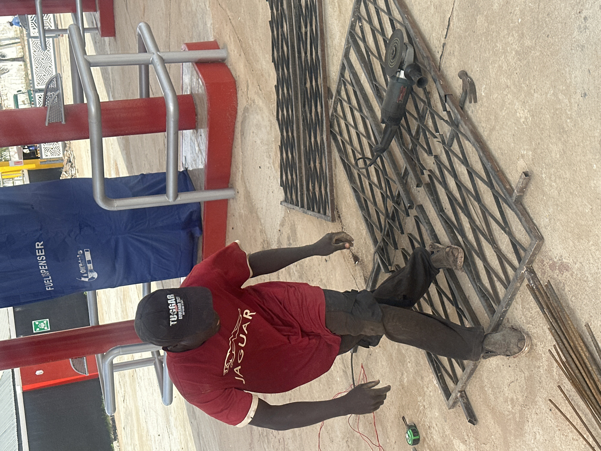 A Black man is working on metal grating at a gas station in Barakin Lerawi, Nigeria. He is wearing a red t-shirt with a Jaguar logo and a black baseball cap with white lettering that reads "TUGGAR GOVERNOR 2023". He is also wearing dark pants and work boots. In front of him are two sections of metal grating, a hammer, a measuring tape, and an angle grinder. Behind him, a blue tarp with "FUEL DISPENSER" printed on it is draped over a structure. Red pillars and silver railings are visible in the background, suggesting an outdoor, possibly commercial, setting.