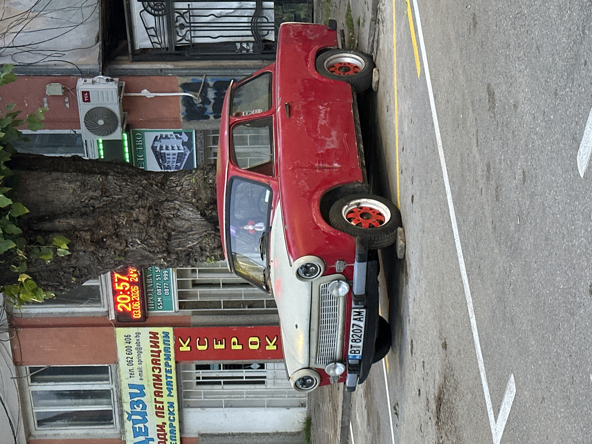 The image depicts an urban street scene in Veliko Tarnovo, Bulgaria, during the evening. A vintage red and white two-door car, possibly a Trabant, is prominently parked on the side of the street. The car has a white front grille and bumper, with red and white spoked wheels. Its Bulgarian license plate reads "BT 8207 AM".
The background features a building with several commercial establishments. An outdoor LED sign displays the time "20:57" and the date "03.06.2026", along with "24°". Below this, a sign advertises "ПРОДАВАА GSM 0877 51 5F 0877 999". Another sign, with yellow lettering, reads "KCEPOK" (likely "ксерокс," meaning xerox or copy shop). A larger yellow sign below it displays "ДЕЙЗИ" with a phone number "062 600 406" and email "spring@abv.bg," advertising "ОДИ, ЛЕГАЛИЗАЦИИ ЕЛАРСКИ МАТЕРИАЛИ" (likely translations, legalizations, and office supplies). A TCL air conditioning unit is mounted on the building wall. A large tree with a textured trunk occupies the left side of the frame. The street has white and yellow painted lines, and the overall lighting suggests it is either dusk or an overcast evening. No people are visible.