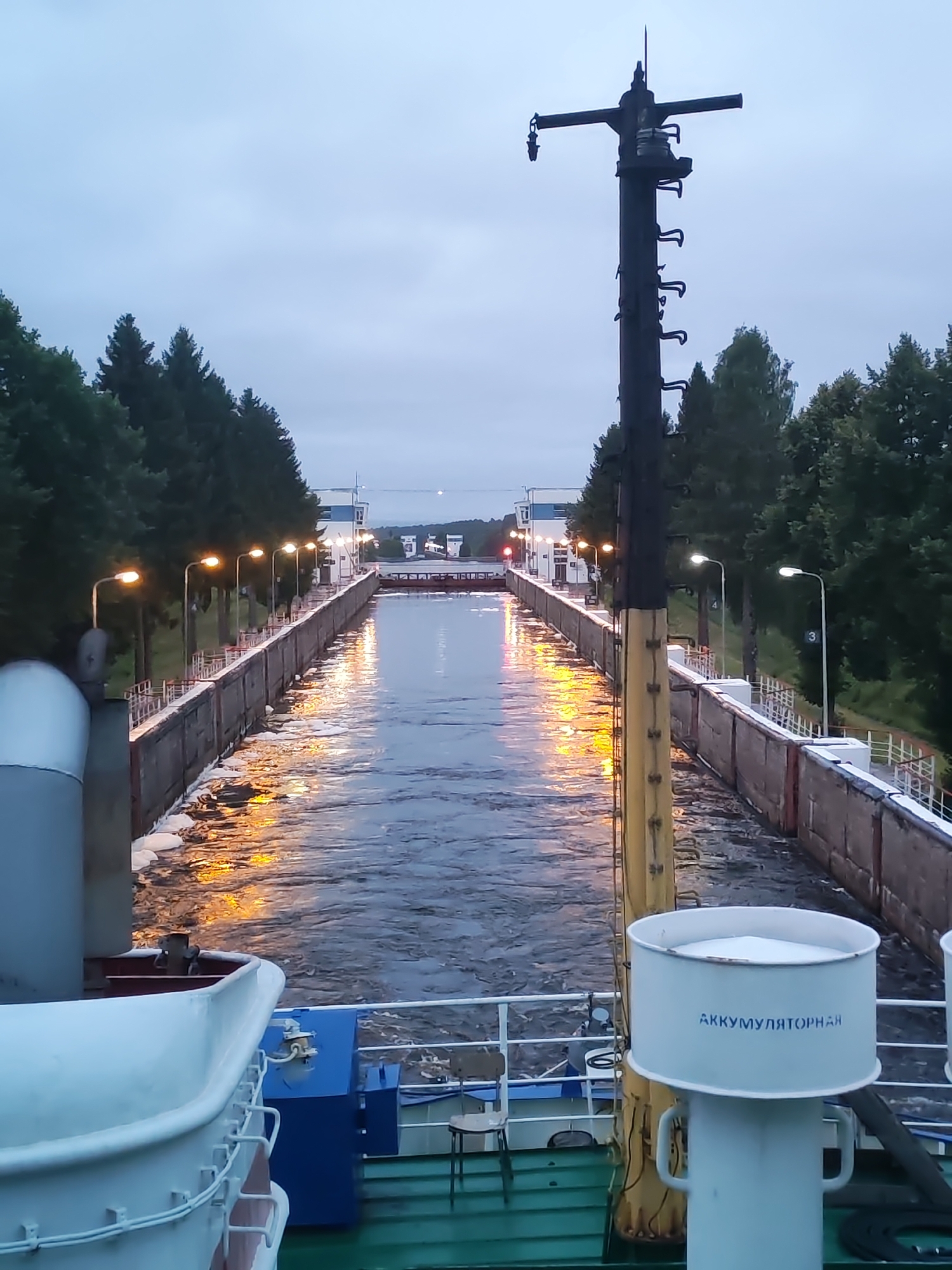 The image depicts a view from a vessel entering a ship canal lock at dusk. The lock walls are lined with trees on either side and illuminated by streetlights, casting reflections on the water. A large metal utility pole stands prominently in the foreground to the right, with a label that reads "АККУМУЛЯТОРНАЯ" (AKKUMULYATORNAYA) on a cylindrical object. The sky is overcast. The scene appears to be in Alekseevskoe, Russia, a town known for its port facilities.