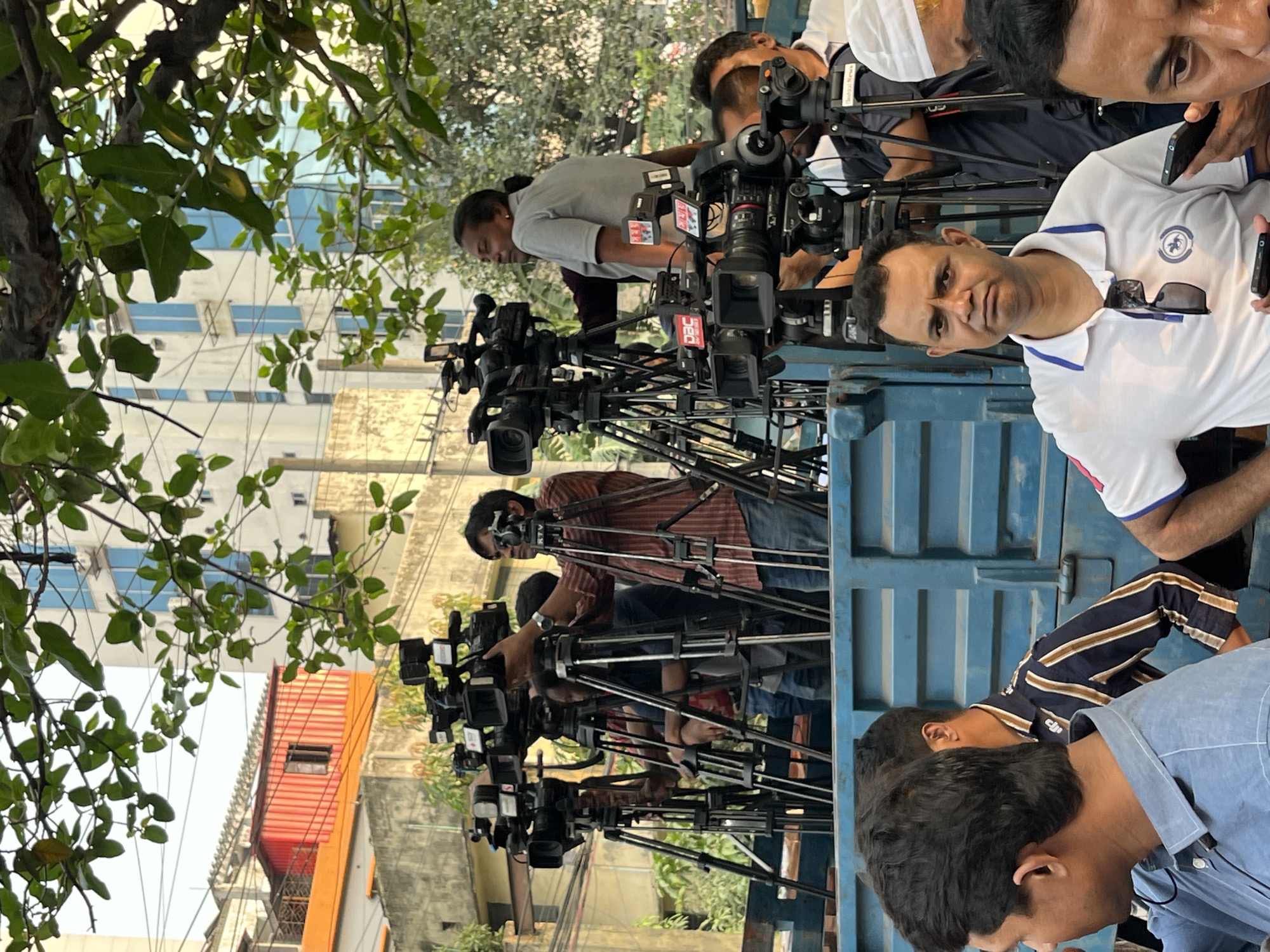 A group of cameramen with professional video cameras on tripods is assembled in the back of a blue truck. The cameramen are of South Asian descent. The cameras have branding on them; specifically, the logo "DAC" is visible on some cameras. People are standing near the cameras.
Outside the truck, there is a street with trees and buildings. The buildings include a tall, blue and white building and a red and orange building. The presence of cameras suggests a media event is about to occur, or has already occurred. Given the branding on the cameras, it is likely this scene is in Dhaka, Bangladesh.