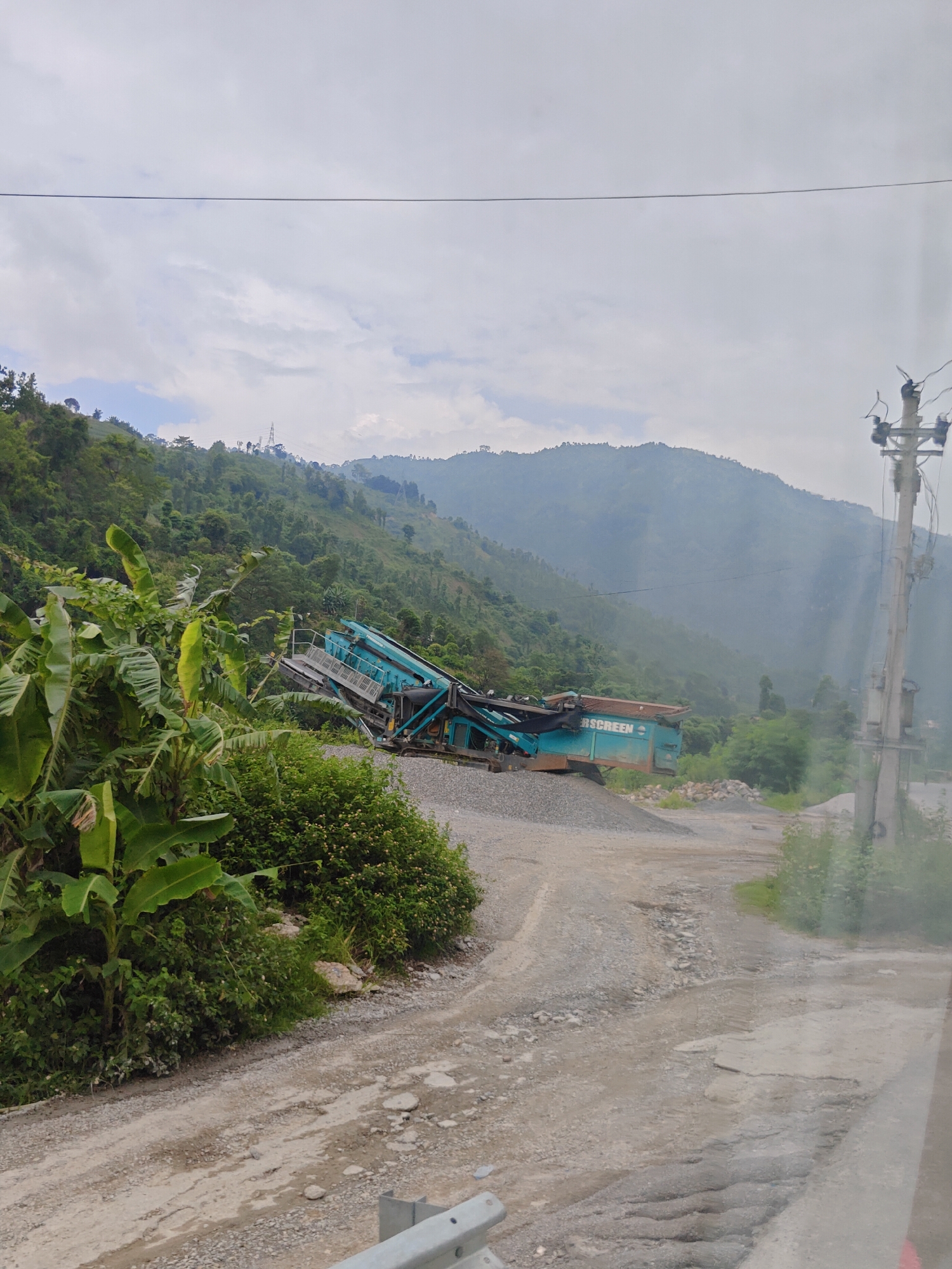 This image captures a rural industrial scene in Thakre, Nepal. In the mid-ground, a large, teal-colored rock crushing machine, possibly a Powerscreen brand, is positioned on a mound of gravel. The machine is angled, suggesting it is in operation or recently used. To the left of the machine, lush green banana plants and other vegetation dominate the foreground, indicating a tropical or subtropical environment. The background features a steep, verdant hillside covered in trees, ascending to misty, mountainous terrain under an overcast sky. A dirt road with scattered rocks and puddles leads through the scene. A utility pole with wires is visible on the right side of the image, suggesting the presence of infrastructure in the otherwise natural landscape. There are no people visible in the frame.
