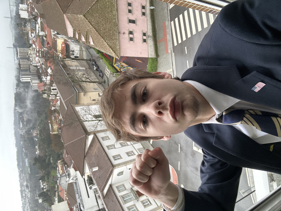 The image presents a young Caucasian male captured in a selfie-style photograph from a high vantage point overlooking the city of Vila Real, Portugal, under an overcast sky. The male subject has light brown hair and a clean-shaven face. He is dressed in a dark blue suit jacket, a white collared shirt, and a dark blue and gold striped tie. A small American flag lapel pin is affixed to his left lapel. His right arm is bent, and his hand is clenched into a fist, raised near his right shoulder. A gold-colored beaded bracelet is visible on his right wrist. He maintains a direct gaze towards the camera with a serious expression, lips slightly pursed.
The background reveals an urban landscape. Immediately below the subject, a wet asphalt street features a white striped crosswalk. Adjacent to the street, a small children's playground with multi-colored structures is visible. The midground consists of residential buildings with light-colored facades and traditional terra cotta tiled roofs, lining narrow streets where parked cars are present. Further into the background, the city expands across hillsides, showcasing a mix of older architecture and taller, more modern apartment buildings. In the far distance, a large concrete bridge, identifiable as the Viaduto do Corgo, spans a valley. The sky is uniformly grey and overcast, with visible mist or low clouds partially obscuring distant hills.