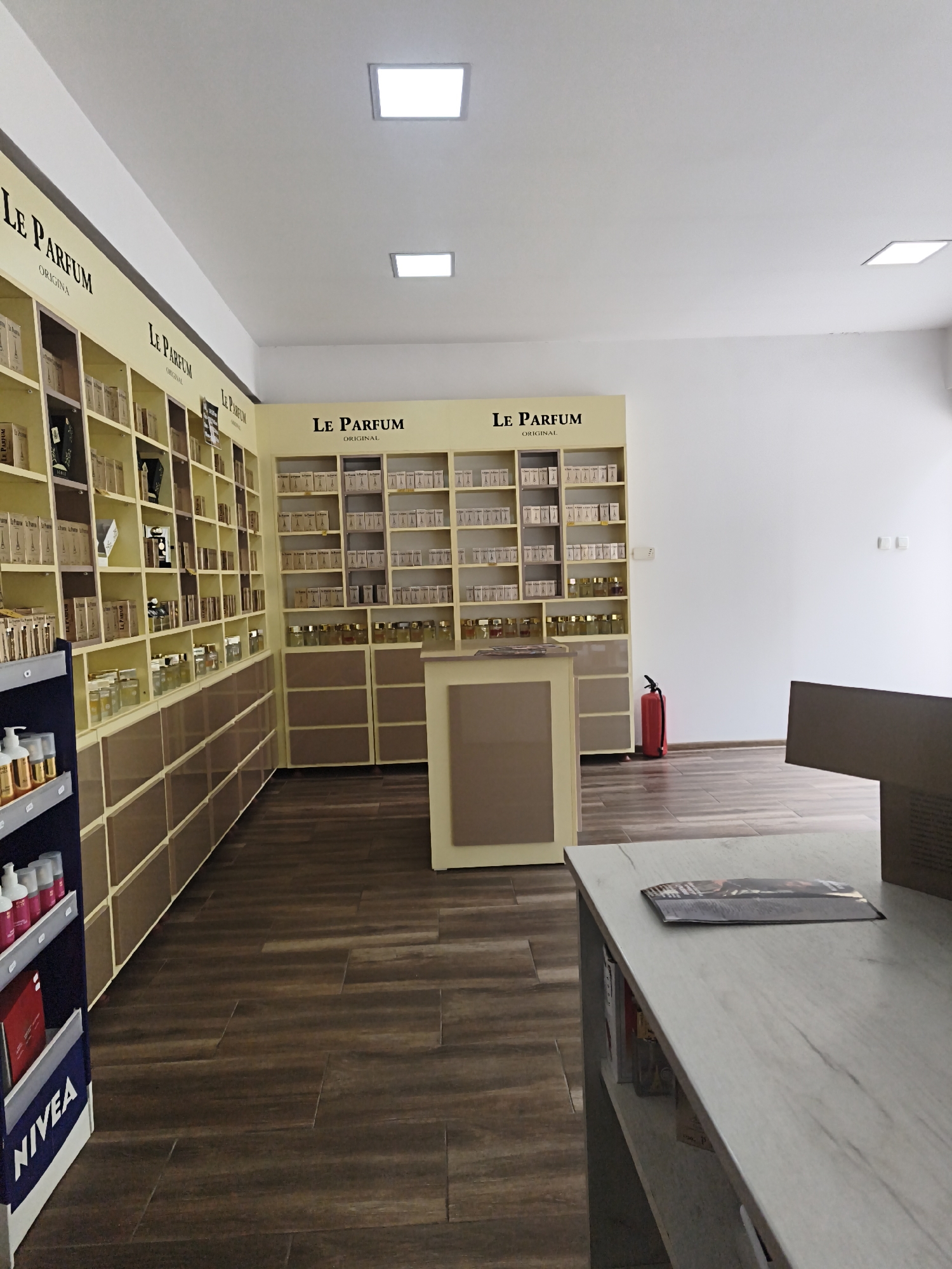 The image shows the interior of a perfume shop. The walls are white, and the ceiling has recessed lighting fixtures. The floor is made of dark wood-like tiles arranged in a horizontal pattern.
The shop features multiple shelves filled with perfume bottles and boxes. The brand "Le Parfum" is prominently displayed in large black lettering on several of the shelving units. There are also lower cabinets with drawers along some of the walls. A central counter unit with a lighter colored top is present in the middle of the room.
On the left side of the image, a dark blue display rack contains bottles of various products and a NIVEA branded bin holding magazines. A red fire extinguisher is visible on the right side of the room near a wall. A brochure or magazine is placed on the counter in the foreground. There are no people visible in the image. The overall impression is of a clean and organized retail space. The location is identified as Harmanli, Bulgaria.