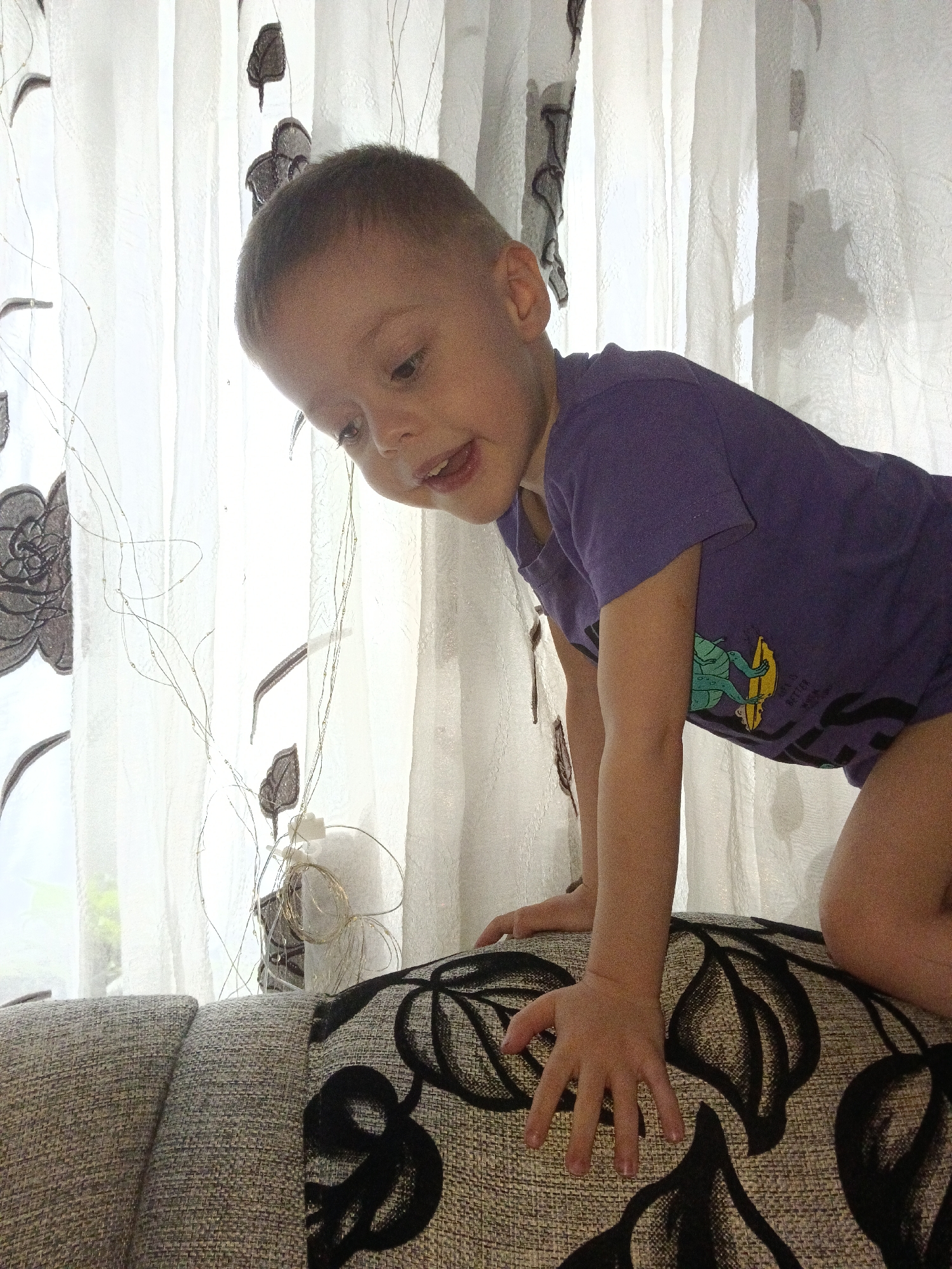 A young child wearing a purple shirt is positioned on a patterned ottoman or cushion. The child is leaning forward with their hands resting on the surface, gazing downwards. Behind the child are light-colored curtains with dark floral patterns. The location is Chaikovsky, Russia.