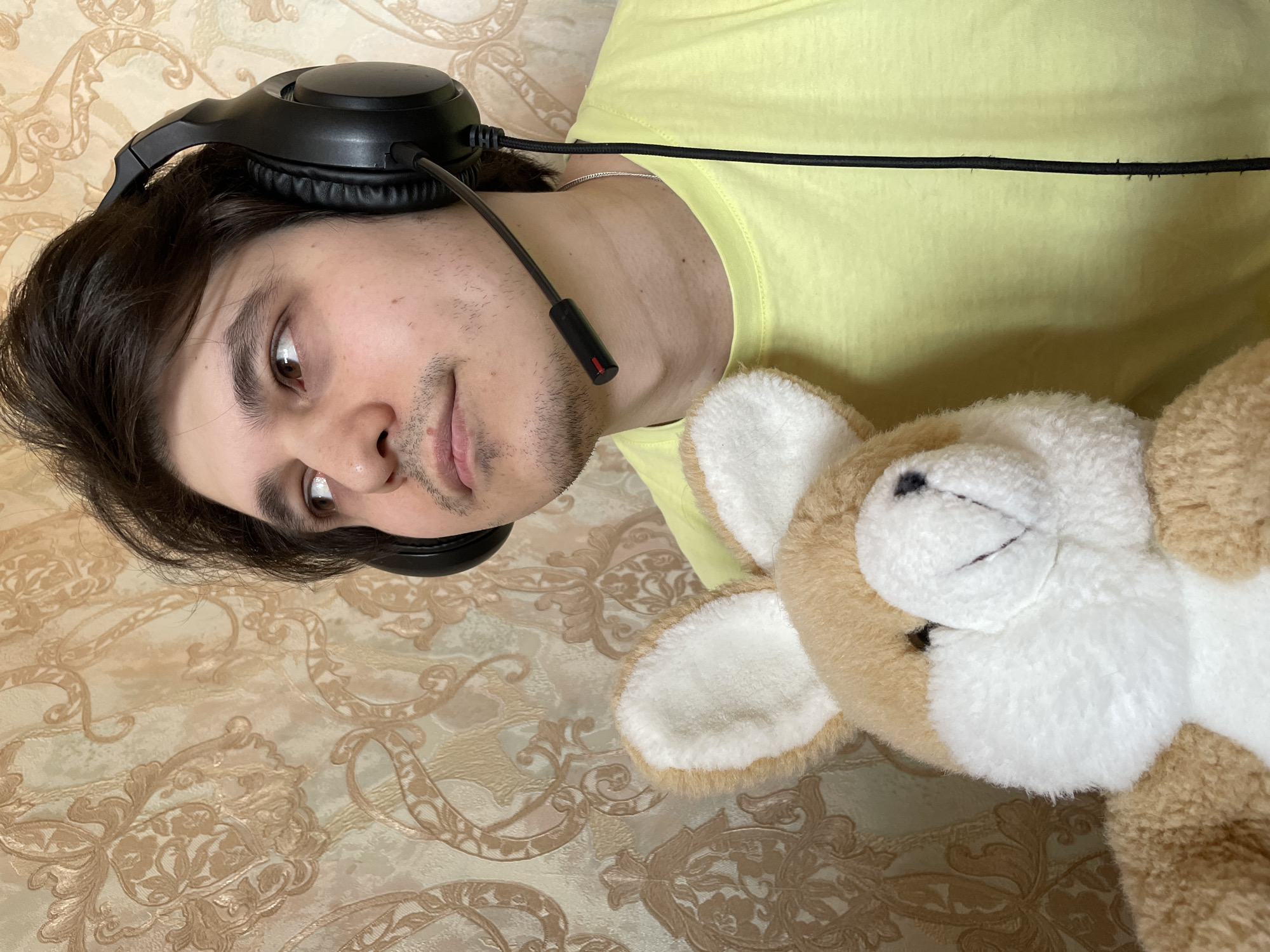 A person lies on a surface patterned with a floral design. The person is wearing a yellow t-shirt and a black headset with a microphone. A plush toy rabbit is located beside the person. The location of the scene is unknown and cannot be determined from the image.