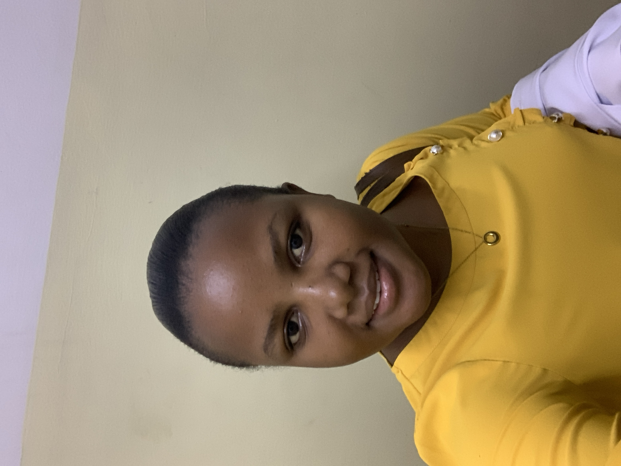 A young woman is looking at the camera, smiling. She is wearing a yellow top with a gold necklace. The background is a plain light yellow wall. It is impossible to determine if this image was taken in the city of Dar es-Salaam, Tanzania.