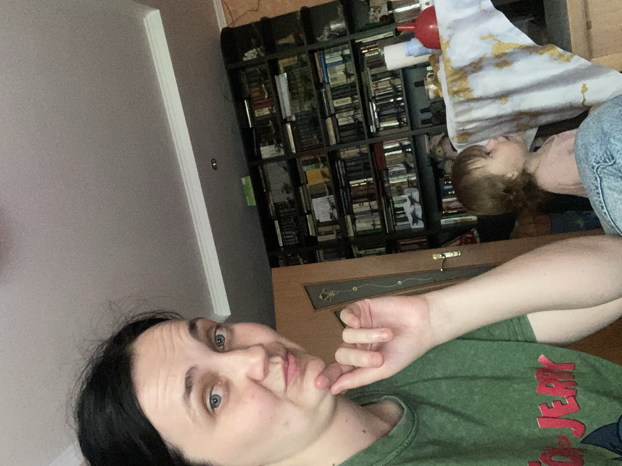 The media file contains a selfie of a person with dark hair and light blue eyes, wearing a green t-shirt. The person is looking at the camera with their hand resting on their chin, suggesting a thoughtful or pensive expression. In the background, a young child with brown hair and a pink shirt is visible, lying down and looking upwards. Behind the child is a bookshelf filled with books. There is also a piece of fabric with a gold pattern draped over the bookshelf. The scene appears to be indoors, possibly in a living room or bedroom, with a wall and some furniture visible. The overall mood seems relaxed and casual. There is visible text on the green t-shirt which reads "JERK" in large red letters.