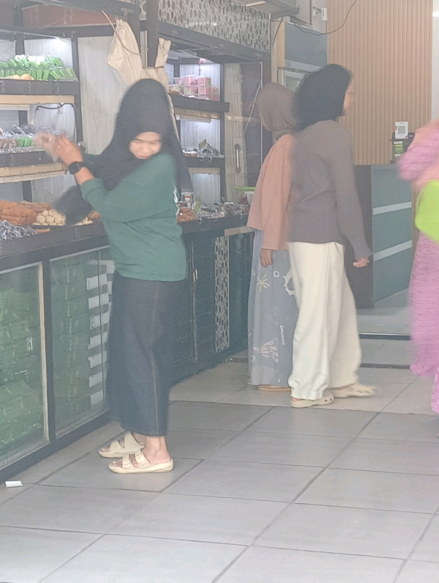 A Muslim woman wearing a black hijab and a green long-sleeved shirt over a dark skirt, and beige sandals, stands to the left of the frame. She is reaching out and holding a clear plastic bag. Behind her, to the right, two other women are visible. One wears a peach-colored hijab and a blue patterned skirt, and the other wears a dark brown jacket and loose-fitting white pants. A partially visible figure in a bright pink and green outfit stands further to the right, partially obscured by the other individuals. The scene appears to be inside a market or shop, with shelves stocked with various food items, including what looks like pastries and fried snacks, visible in the background. The flooring is tiled. The overall lighting suggests an indoor setting, possibly a retail establishment. The location is identified as Bandar Lampung, Indonesia.