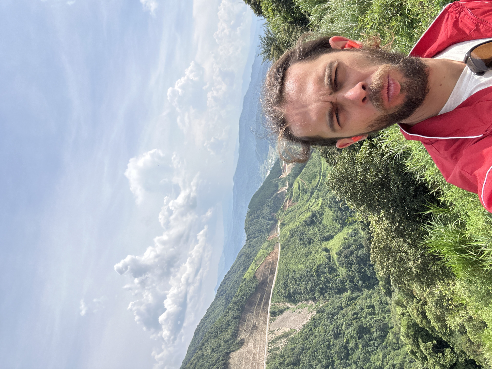 A selfie-style portrait captures a man outdoors against a vast mountainous landscape, with the image rotated 90 degrees clockwise from its natural orientation. The man, appearing to be a white male with a beard, mustache, and medium-length dark hair, is prominently featured in the foreground. He is dressed in a red jacket with white trim over a white t-shirt, and distinctive orange earplugs are visible in both ears. Sunglasses are perched on his head or collar. His eyes are squinted or closed, and his lips are slightly pursed, suggesting a reaction to bright sunlight or a playful expression.
Behind the man, a panoramic view unfolds, showcasing lush green mountains and deep valleys under a bright sky adorned with scattered white clouds. The scene suggests a high elevation, offering an expansive vista. A notable feature in the mid-ground is a long, cleared path or road-like structure cutting through the densely forested mountain slopes, possibly indicating a construction project or a past geological event. The overall impression is of a vibrant, sunny day in a serene and grand natural environment. The location is Muong Than, Vietnam. No text is visible in the image.