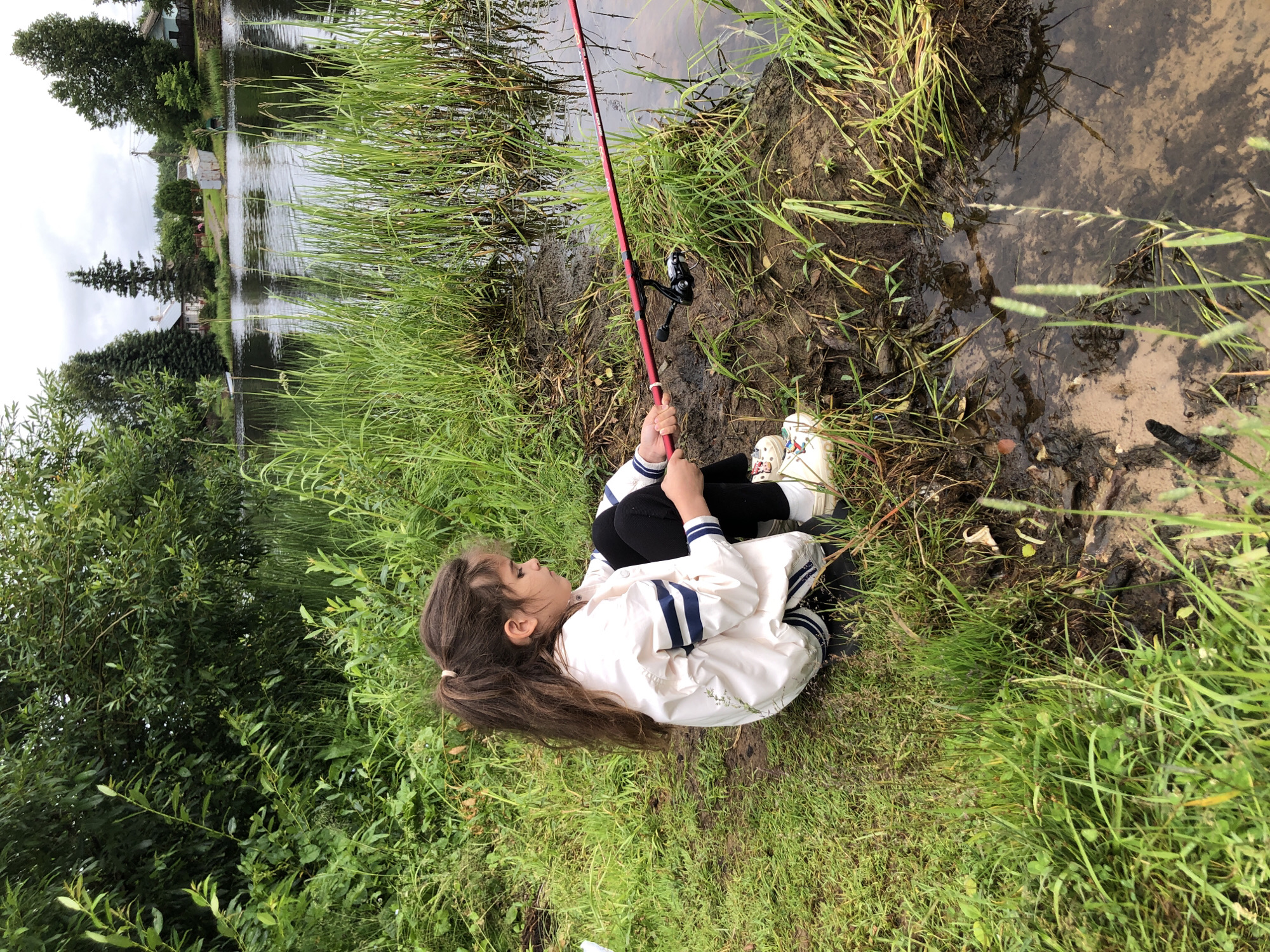 This media file depicts a young girl sitting on the grassy bank of a body of water, holding a fishing rod. The girl is wearing a white jacket with blue stripes on the sleeves and collar, black pants, and white sneakers. She has long, dark hair tied back in a ponytail. Her gaze is directed towards the water, and she appears focused on her fishing.
The scene is a natural environment with lush green grass and tall reeds growing along the water's edge. The water itself is calm, reflecting the overcast sky. In the background, across the water, several houses and trees are visible, suggesting a semi-rural or residential setting. The weather appears to be cloudy, contributing to a muted light.
The activity is fishing, with the girl actively engaged in the pursuit. The fishing rod is red with a black reel. The ground where she is sitting is covered in grass, with patches of dark, wet soil near the water.
Notable details include the girl's posture, indicating concentration, and the overall peacefulness of the scene. The presence of the houses in the background provides context about the location. No visible text is present in the image. The time of day is indeterminate due to the overcast conditions, but it appears to be daylight.