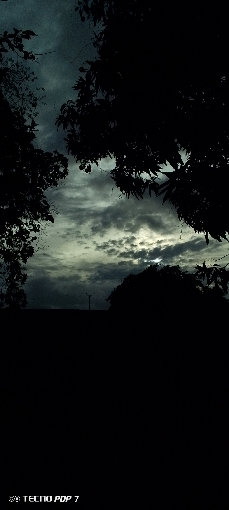 This image is a vertical shot of a darkened sky with clouds and silhouettes of trees. The sun is partially visible through a break in the clouds, casting a faint glow. In the foreground, there is a dark silhouette of a wall or building, with a lamppost visible on top. The bottom of the image contains a watermark that reads "TECNO POP 7". The location is specified as Altagracia, Venezuela.