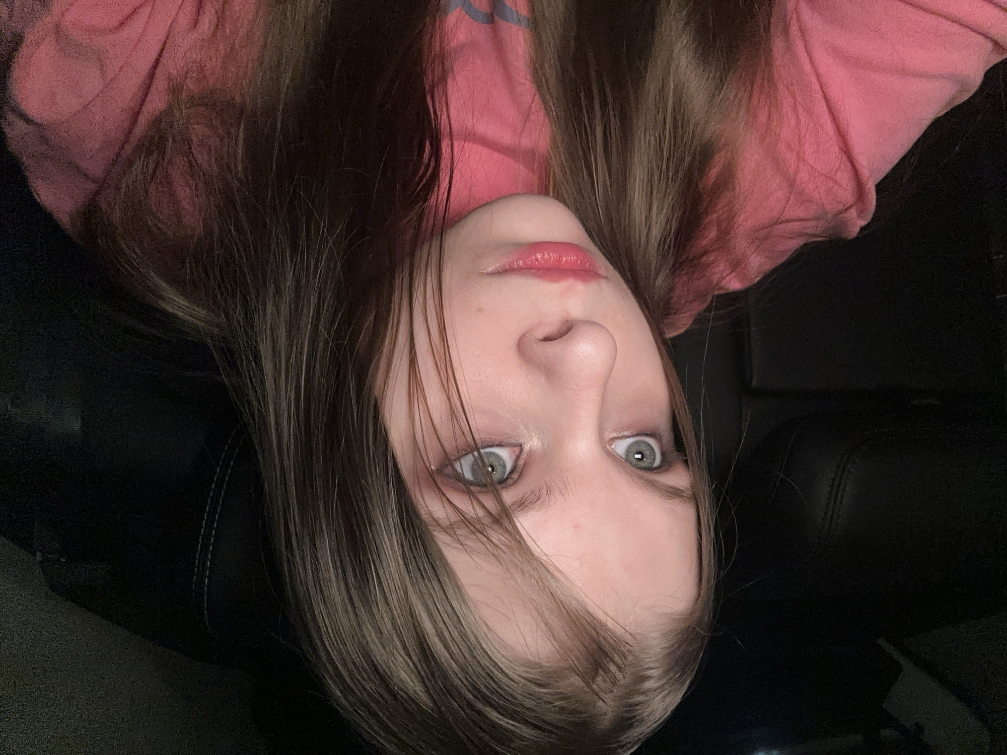 A young woman with long brown hair and green eyes is shown in an upside-down selfie. She is wearing a pink shirt. The background appears to be the interior of a dark vehicle, possibly a car. There is no indication of the image's location.