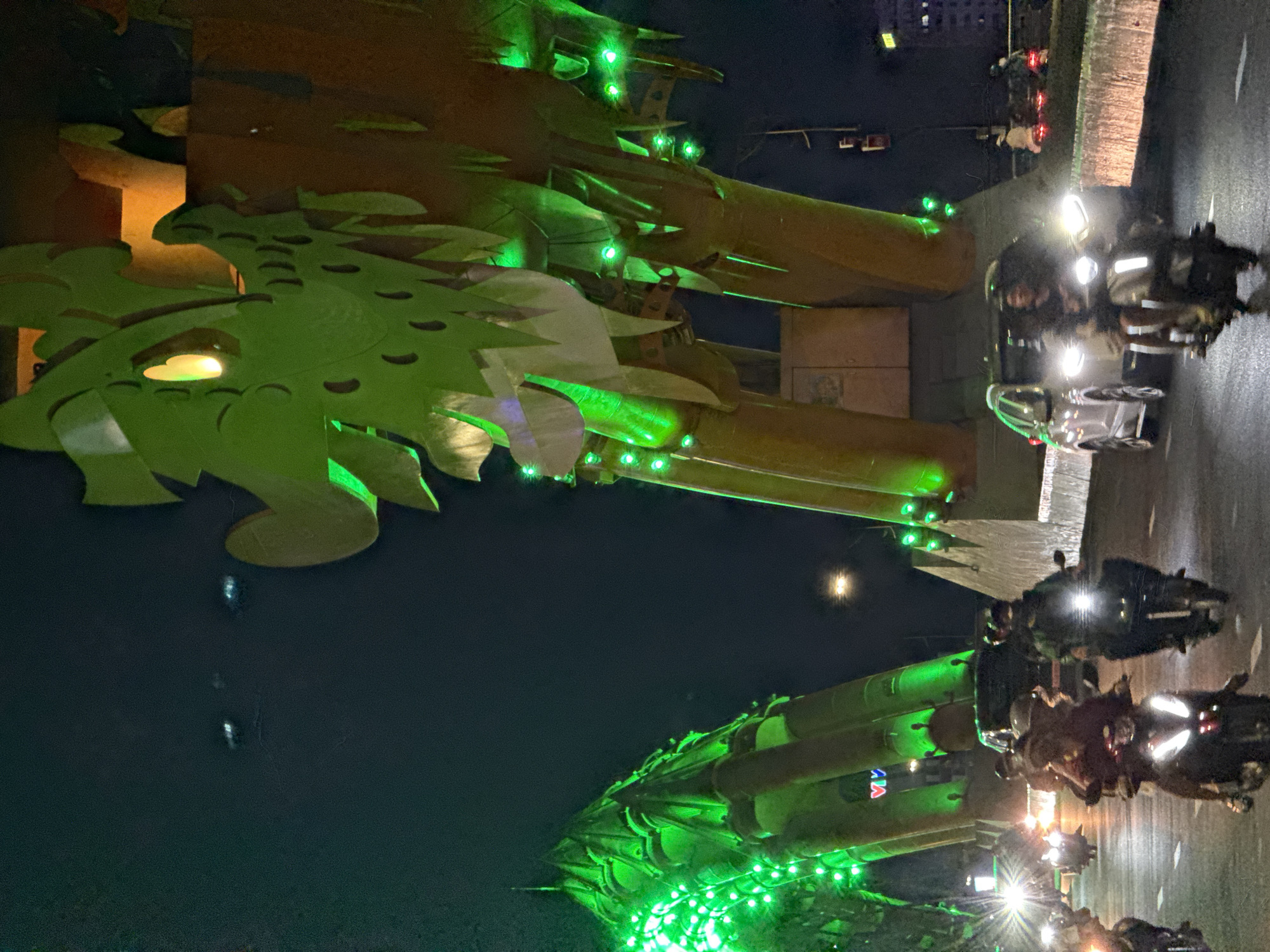 The image presents a vibrant night scene in Đà Nẵng, Vietnam, centered around the iconic Dragon Bridge. The bridge's large, stylized dragon structure, in a golden-brown or metallic tone, is prominently illuminated with numerous bright green lights, particularly along its scales and eyes, creating a striking spectacle against the dark sky.
A multi-lane road occupies the right side of the frame, bustling with activity. Numerous motorcycles and at least one car are visible, their headlights and taillights glowing brightly as they traverse the road. Several people are seen on the motorcycles, wearing helmets, indicating a typical evening commute or drive.
The sky is dark, signifying nighttime, with a clear, bright circular light source, likely the moon, visible in the upper right. Distant buildings are dimly lit in the background. On the side of one of the bridge's supporting structures, red and blue letters spelling "DA" are visible, along with other indistinct characters, likely part of a city or promotional sign. The scene captures the lively urban atmosphere of Đà Nẵng at night, with the illuminated Dragon Bridge serving as a magnificent landmark and a focal point of city life.
