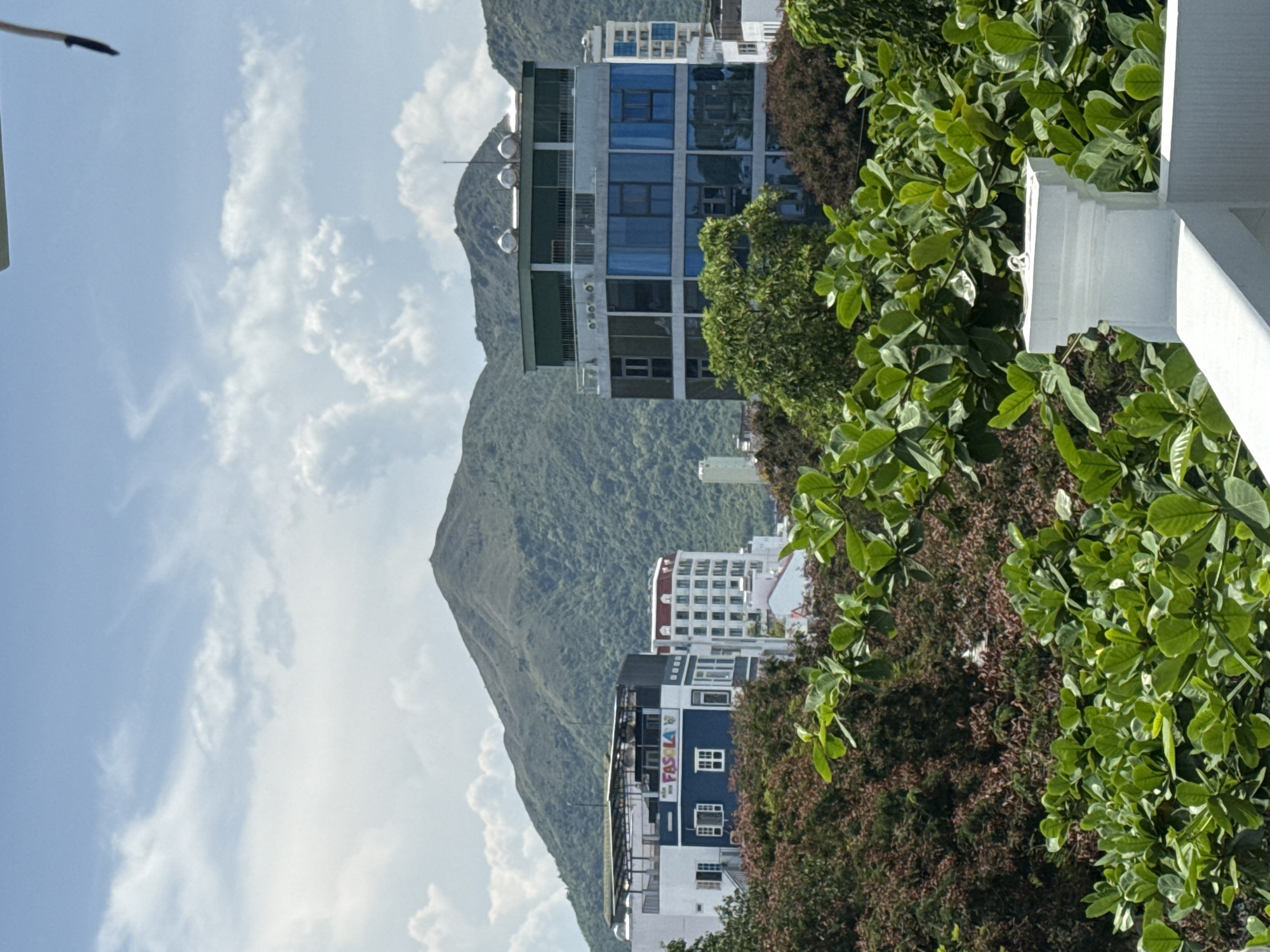 This is a photograph taken in Bac Nha Trang Ward, Vietnam, capturing a view of buildings against a backdrop of lush green mountains under a partly cloudy sky. In the foreground, vibrant green foliage from trees and bushes obscures the lower portion of the scene. A white, ornate railing or structure is visible on the right side of the frame, leading the eye towards the cityscape.
Several buildings are prominent in the midground. To the left, a white building with blue accents and visible windows stands, with a sign that reads "FASOLA" with colorful lettering. Adjacent to it, to the right, is a taller, modern building featuring a facade of blue glass windows on its upper levels and what appears to be a green awning or roof structure. Further to the right, another white building with arched windows and a red roof is visible. In the distance, hints of other high-rise buildings can be seen.
The background is dominated by a large, verdant mountain range, its slopes covered in dense vegetation. The sky above is a pale blue, dotted with scattered white clouds, some of which are tinged with gray, suggesting the possibility of recent or approaching rain. The overall impression is one of a tropical or subtropical urban environment nestled within a natural landscape. No people or specific actions are discernible.