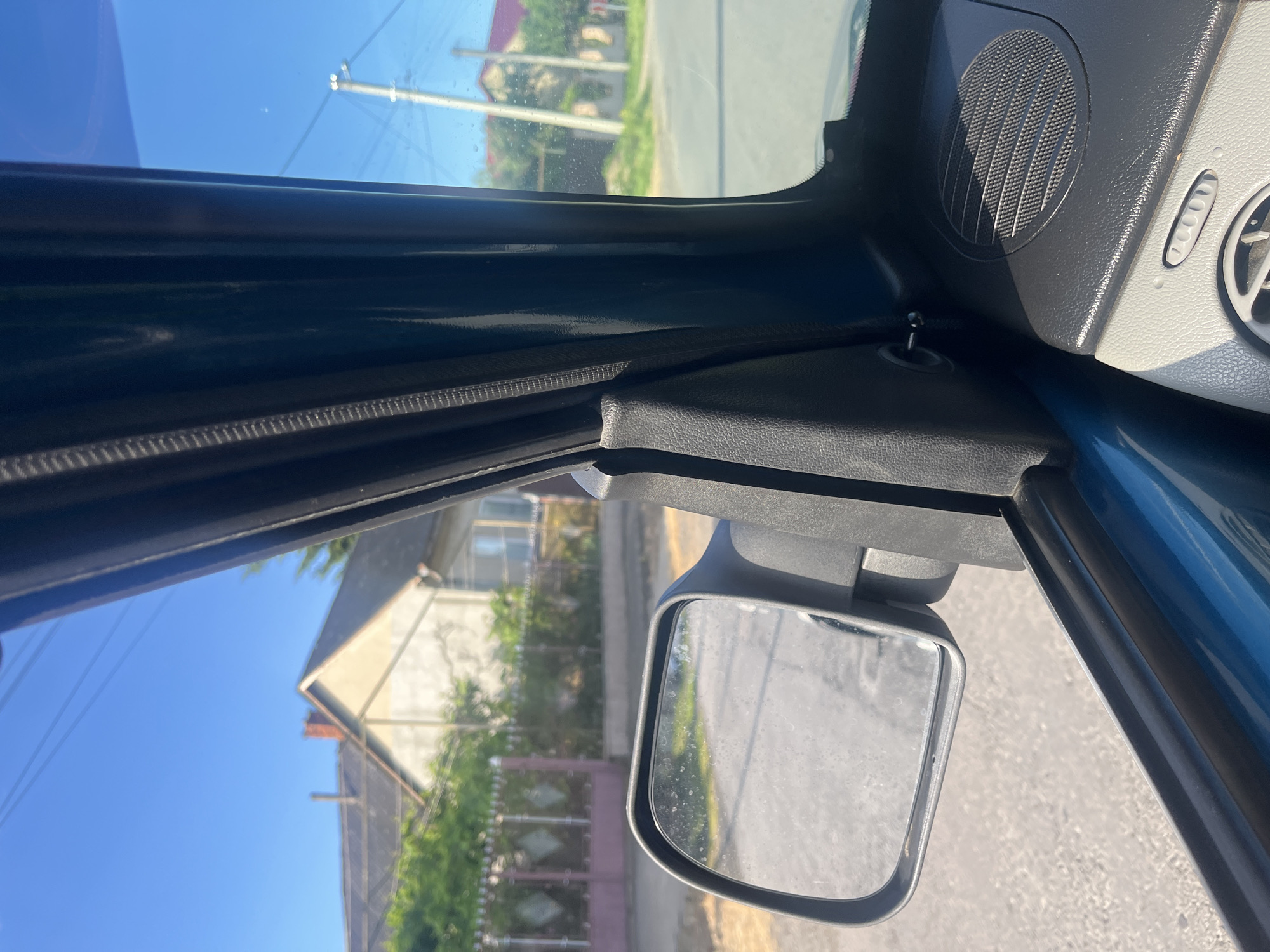 The image is taken from inside a car, looking out the passenger side window towards the side mirror and the surrounding environment. The scene depicts a sunny day in a residential area, likely in Tarnivtsi, Ukraine.
In the foreground, the car's interior is visible, including a portion of the dashboard with a speaker grille and a vent. The side mirror is prominently displayed, reflecting a blurred image of a road and possibly the sky. The window frame of the car, made of dark plastic, runs vertically along the right side of the image.
Outside the car, through the side window and reflected in the mirror, a residential street can be seen. Houses with pitched roofs, some with visible chimneys, are situated along the street. Green trees and bushes line the properties. A decorative fence with a geometric pattern is visible in front of one of the houses. Electrical poles and wires are present in the background, typical of a suburban or rural setting. The sky is a clear, bright blue, indicating good weather conditions.
There are no people or animals visible in the image. The focus is on the view from the car and the details of the car's interior. The overall impression is one of a casual drive through a quiet neighborhood.