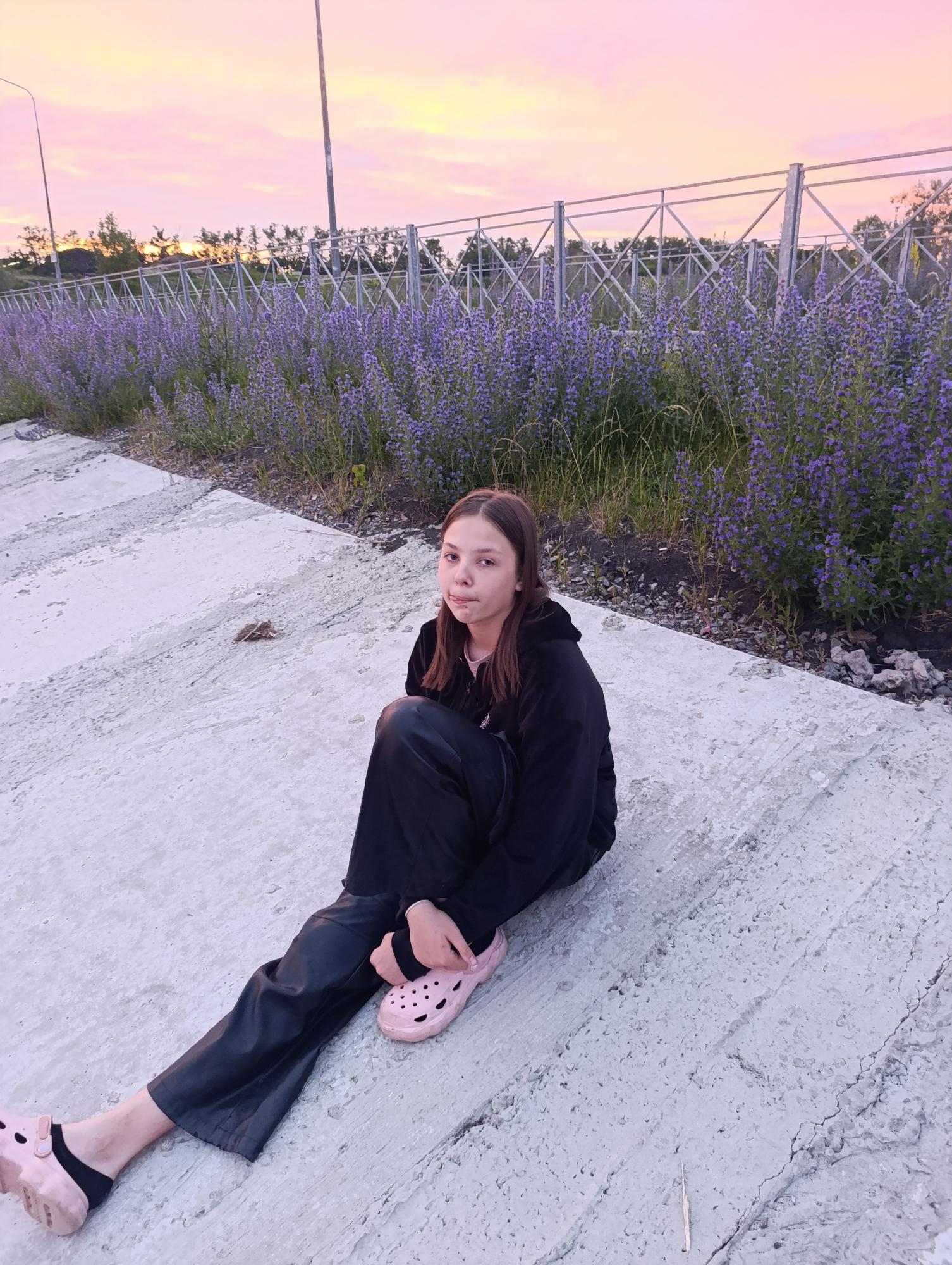 A young person with brown hair is sitting on a concrete embankment, looking at the camera with a neutral expression. They are wearing a black hoodie and black pants, with pink clogs and black socks. Behind them, a line of purple flowers runs alongside a metal fence. The sky is a soft pink and orange, indicating either sunrise or sunset. The scene is set in a park or an area with landscaping in the city of Penza, Russia.