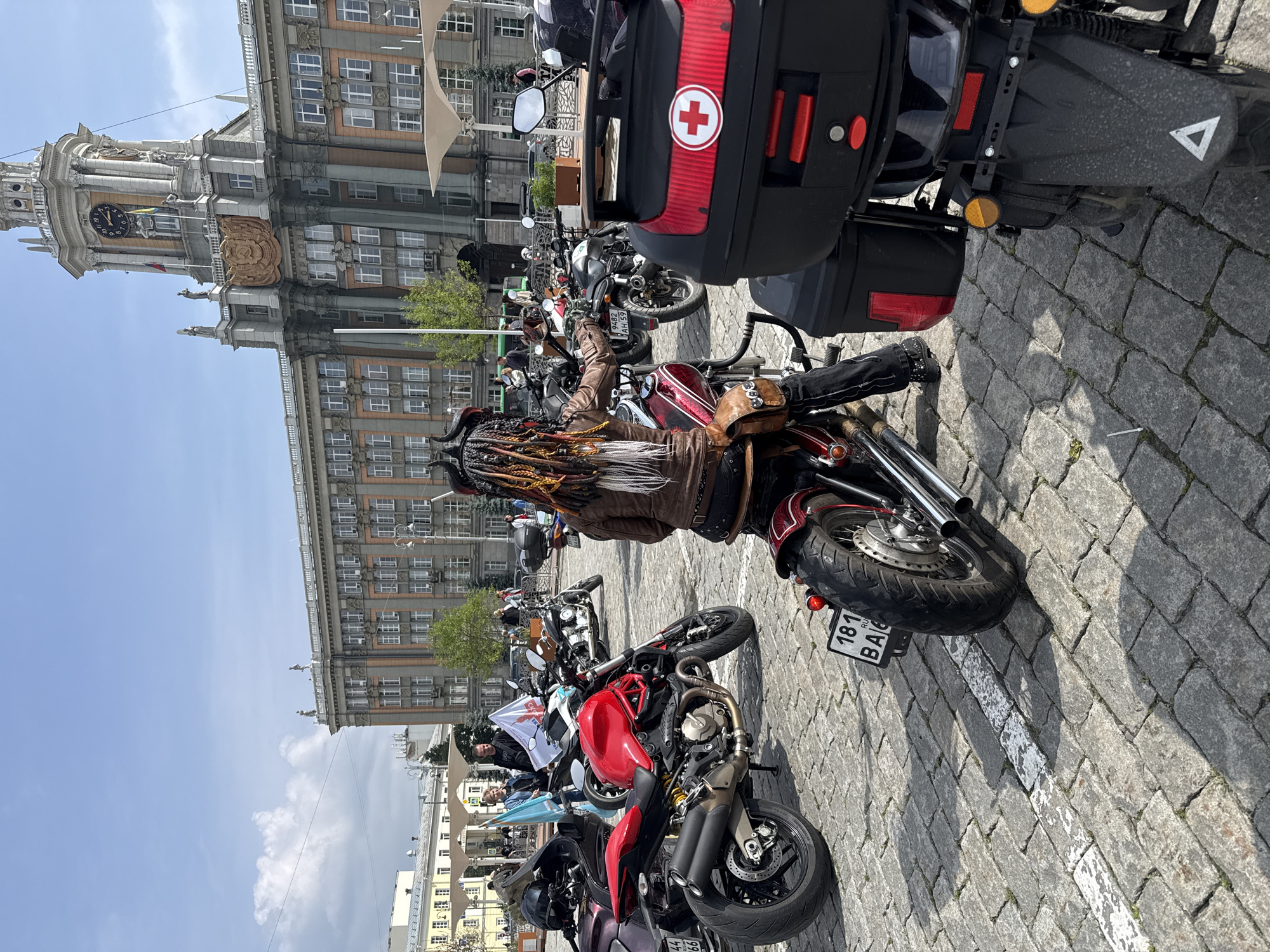 The image depicts an outdoor gathering of motorcyclists on a cobblestone square in Yekaterinburg, Russia, under a clear sky with scattered clouds. The dominant structure in the background is the Government House of Sverdlovsk Oblast, an imposing multi-story building featuring classical architectural elements, a prominent clock tower displaying approximately 11:30, and a large golden crest (the Coat of Arms of Sverdlovsk Oblast) below the clock face. Flags are visible flying from the tower.
In the immediate foreground, facing away from the camera, a person with distinctive long, multi-colored dreadlocks and a horned headpiece is seated on a dark red custom cruiser motorcycle. The person wears a brown leather jacket and dark trousers. The motorcycle has custom exhaust pipes, a wide rear tire, chrome accents, and a brown leather saddlebag. Its Russian license plate reads "181 RU BA6". To the right, the rear of a black motorcycle is visible, equipped with a large black top case featuring a red reflective strip and a white circular decal with a red cross symbol.
The square is paved with grey cobblestones, marked by a faded white line. Numerous other motorcycles of various types are parked throughout the midground, including a bright red Ducati Monster-style motorcycle prominently positioned in the left-midground. Several individuals are visible standing amongst the bikes; two people, a woman in a light blue top and a man in a dark t-shirt, are observed near the red Ducati. Flags, including a light blue one and a white one with a red emblem, are displayed.
